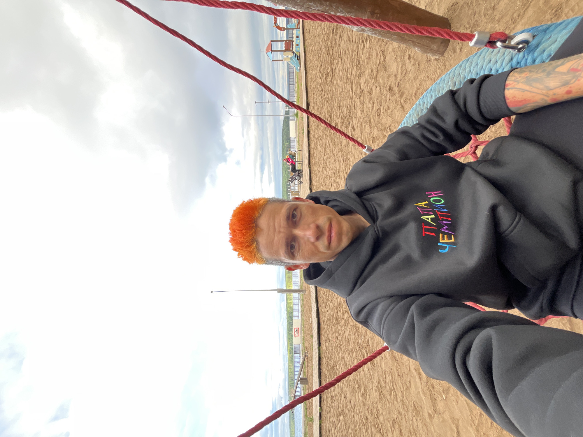 This image displays an outdoor scene in the city of Chaikovsky, Russia, featuring a male individual in a selfie-style photograph. The subject has short, bright orange hair and is wearing a black hooded sweatshirt with "ПАПА ЧЕМПИОН" (Papa Champion) embroidered in colorful Cyrillic letters on the chest. Tattoos are visible on his left forearm. He is seated on a large circular swing with a blue rim, suspended by thick red ropes, in a sandy playground.
In the background, a wooden boardwalk runs parallel to a wide body of water, likely a lake or river, beyond which a dense treeline is visible under an overcast sky. A white fence, bearing a red sign with a white symbol (appearing to be a prohibition or warning symbol), separates the boardwalk from the water. To the right of the frame, a multi-colored playground structure with a slide and climbing elements is present. To the left, two individuals, one with a stroller, are seated on a bench. A tall, slender street light pole stands in the mid-ground.
