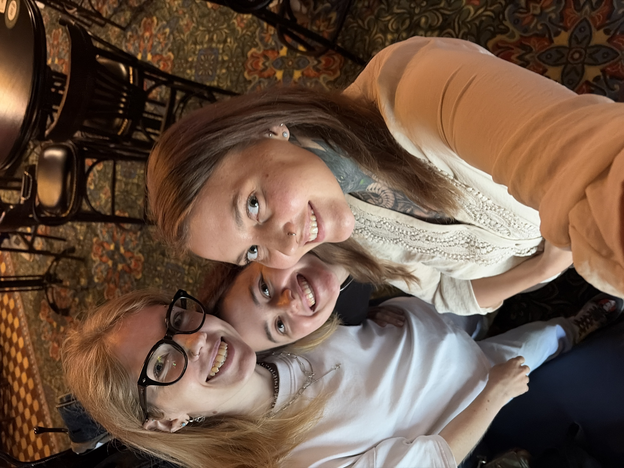 Three individuals, two women and one person whose gender is indeterminate from the provided image, are posing for a selfie in what appears to be a bar or pub. The person on the left, a woman with blonde hair and glasses, is wearing a white sweatshirt and multiple necklaces. The person in the middle, a woman with brown hair, is smiling and wearing a dark top. The person on the right, a woman with brown hair, has tattoos on her neck and arm and is wearing a light-colored, ornate top. They are seated at a table with chairs visible in the background, and the floor is covered with a patterned carpet. The lighting suggests an indoor setting, possibly in the evening. The image does not provide any specific visual cues that would allow for determination of the city as Izhevsk, Russia.