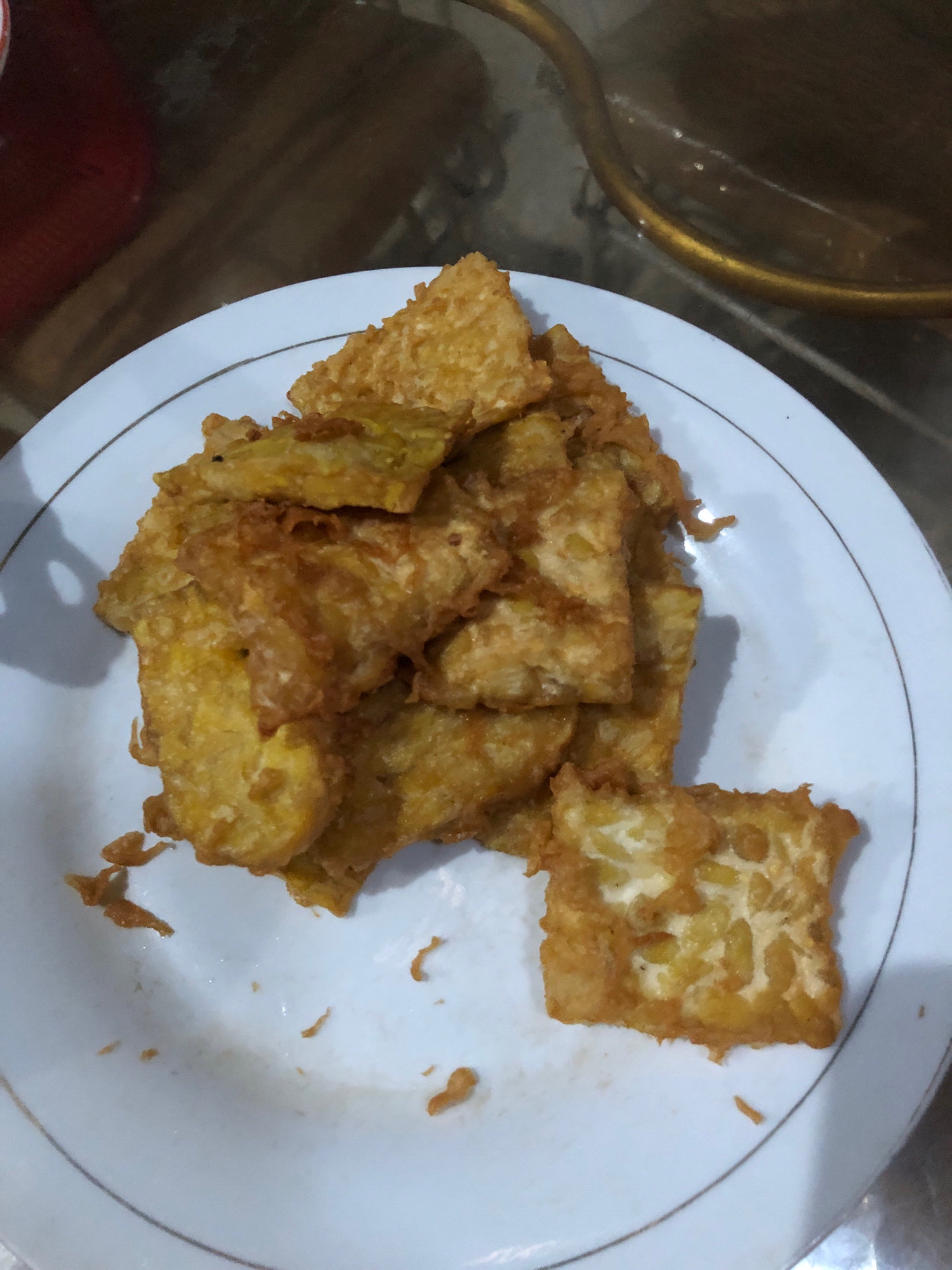 A close-up, overhead shot showcases a white plate with a thin gold rim, piled high with golden-brown fried tempeh. The tempeh pieces are irregularly shaped, some appearing as thin slices, while others are thicker, with visible soy bean texture. A single square piece of tempeh sits apart on the plate, its surface clearly showing the fermented soybean texture. The plate is positioned on a dark, reflective surface, possibly a wooden table, with a blurred red object visible in the upper left corner. A hint of a golden metal object, possibly a utensil or a pot handle, is also visible in the upper right. The lighting suggests indoor, likely domestic, conditions, common in Lhokseumawe, Indonesia, where tempeh is a popular and staple food. The overall scene is a simple, unembellished presentation of a common Indonesian dish.