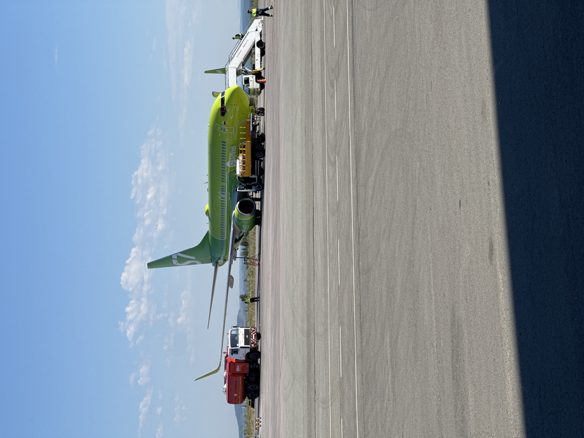An S7 Airlines Boeing 737 aircraft, painted bright green with the S7 logo on its tail, is parked on an airport tarmac under a clear blue sky with scattered white clouds. To the left of the aircraft, a red and white fuel truck is positioned near the wing. A baggage loader, a white vehicle with a yellow loading platform, is situated next to the aircraft's fuselage. A set of mobile stairs is attached to the aircraft's door, with two individuals visible near it. Another person, wearing a high-visibility vest, stands near the fuel truck. The ground is paved with asphalt, marked with white lines. In the background, a hilly landscape can be seen. The image captures a scene at an airport, likely in or near Чульман, Russia, where the aircraft is undergoing servicing or preparation.