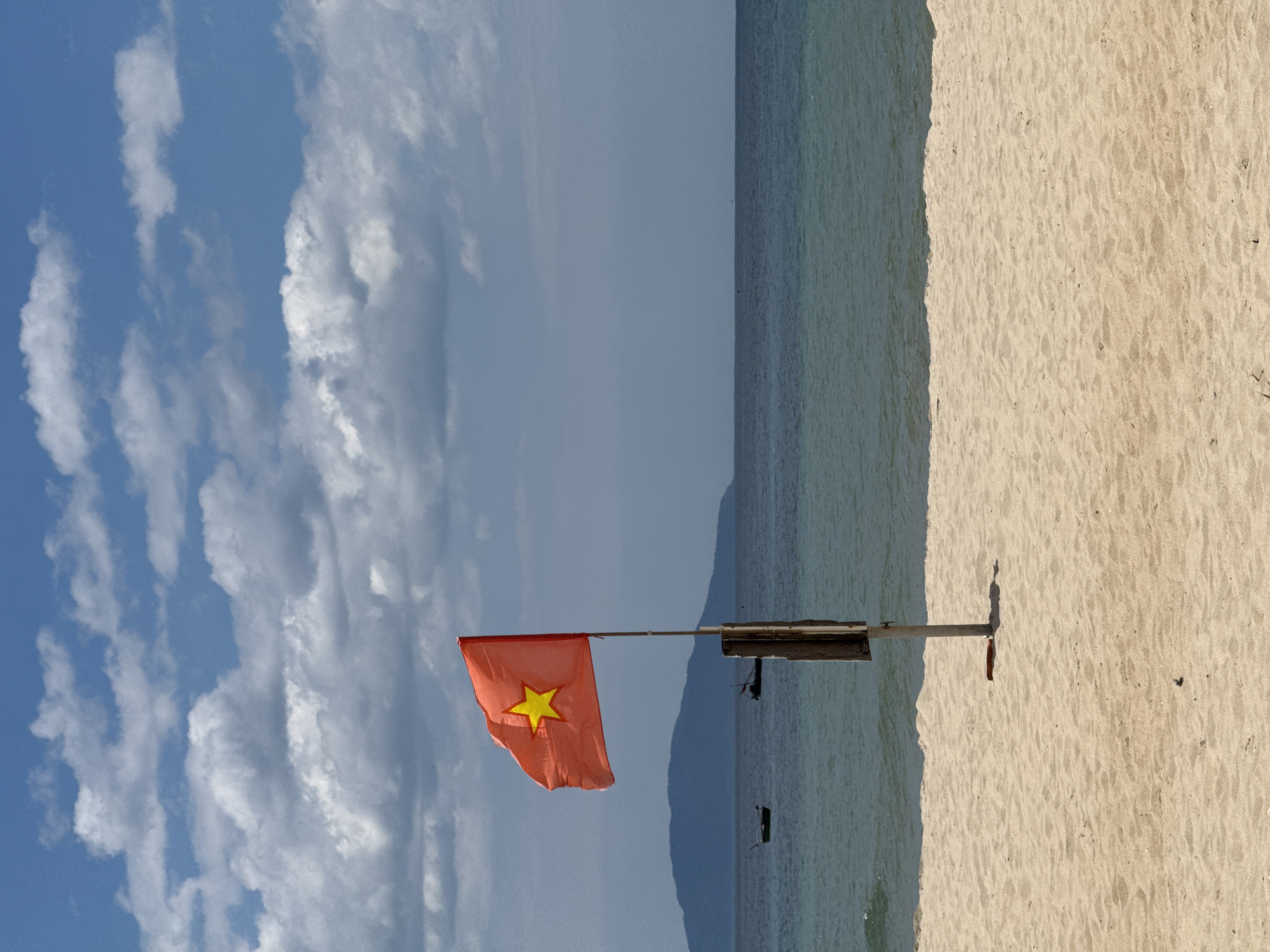 The image depicts a Vietnamese flag on a sandy beach in Đà Nẵng, Vietnam, with the ocean, distant mountains, and a cloudy sky as the backdrop.
In the foreground, a light beige sandy beach occupies the lower third of the frame. The sand is textured with subtle undulations and sparse dark specks, possibly small pebbles or debris. Near the base of the flagpole, a reddish-brown, elongated object rests flat on the sand.
Centrally positioned, an orange-red flag of Vietnam with a large yellow five-pointed star is unfurled and waving leftward, showing creases from the wind. The flag is mounted on a light-colored wooden pole, which is further supported by a darker, secondary wooden post partially visible to its right. The pole is embedded vertically in the sand.
Beyond the beach, the ocean stretches across the midground. The water near the shore is a light turquoise-green, transitioning to a deeper blue further out. Small ripples are visible on the water's surface. Two dark, small boats are positioned in the mid-distance on the water, with one further left and the other slightly right of the flagpole.
In the background, a dark blue-grey mountainous landmass extends along the horizon, partially obscured by a light haze. The sky above is a mix of bright blue and scattered white and grey cumulus clouds. The clouds are denser and darker towards the upper right and horizon, while patches of clear blue sky are visible in the upper left quadrant. No people or animals are visible in the scene.