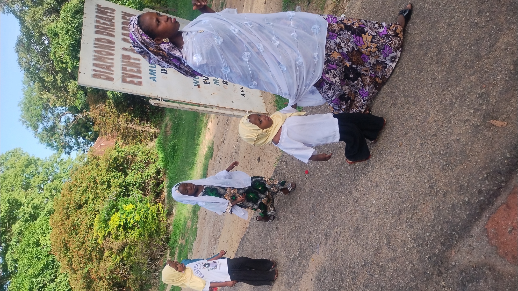 Three girls and a woman are standing on a paved road next to a sign that reads "Diamond Breaks Event & Garden". The woman is wearing a long, floral skirt, a white top, and a head covering. The girls are wearing various dresses and head coverings. Trees are visible in the background. The scene is likely in Jos, Nigeria, based on the sign's location and the clothing worn by the people in the image.