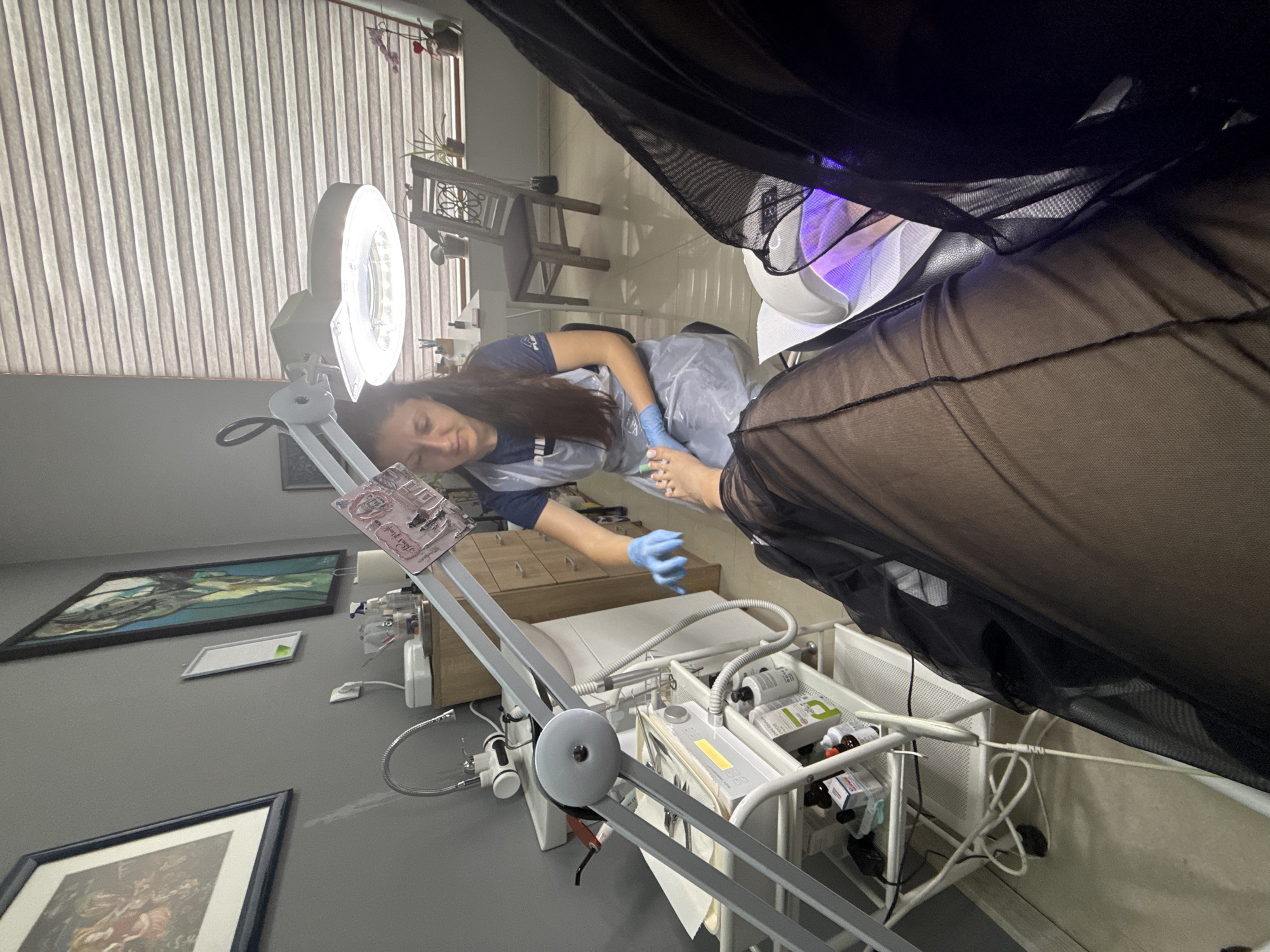 An indoor beauty salon scene, captured from a first-person perspective of a client, shows a female pedicurist performing a pedicure in the city of Unknown, Unknown. The pedicurist, with long dark hair, wears a dark blue t-shirt with a white logo, a clear plastic disposable apron, and blue disposable gloves. She is seated on a low stool, intently applying green nail polish to the client's left foot, which rests on a white support.
The client's legs are visible, covered in sheer black fabric, likely hosiery or a protective wrap. The client's right foot is positioned inside a white UV/LED nail lamp, which emits a purple light for curing nails.
Various professional equipment surrounds the work area. A tall, articulated magnifying lamp with a circular LED light source extends over the client's feet, providing illumination. To the left, a multi-tiered white utility cart holds an array of pedicure instruments, including metal tools, small bottles of solutions, and white disposable coverings. Behind the cart, a small white sink with a flexible chrome faucet is installed next to a light wood cabinet topped with additional cosmetic supplies like spray bottles and cotton rolls.
The room features neutral gray walls. Two framed pictures hang on the left wall: one depicting a classical-style scene and another with an abstract design, possibly a horse's head in green and blue tones. A smaller white framed item is situated between them. In the background, a large window with light-colored horizontal pleated blinds allows diffused natural light into the room. A decorative wooden chair and several small potted plants are arranged on the windowsill. The floor is covered with light-toned tiles, and some electrical cords are visible near the equipment.