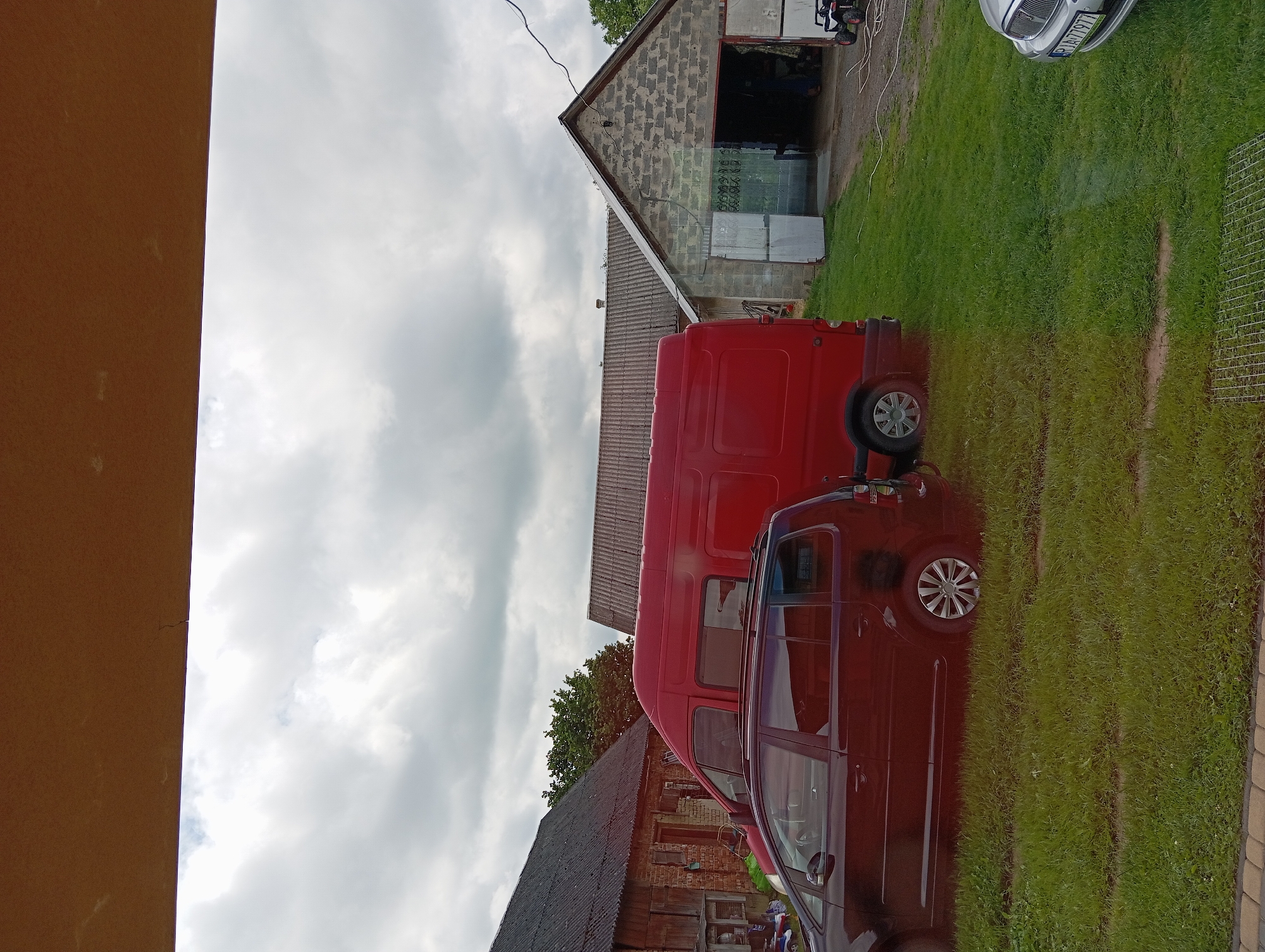 The image depicts an outdoor scene in Hlynytsi, Ukraine. In the foreground, a dark-colored station wagon is parked on a grassy area. Behind it, a bright red van is also parked. To the right, a concrete building with a corrugated roof and two open garage doors is visible. Inside the garage, some objects and a person can be partially seen. Further to the left, a brick building with a dark, corrugated roof is present. The sky above is overcast with clouds. The ground is covered with green grass.