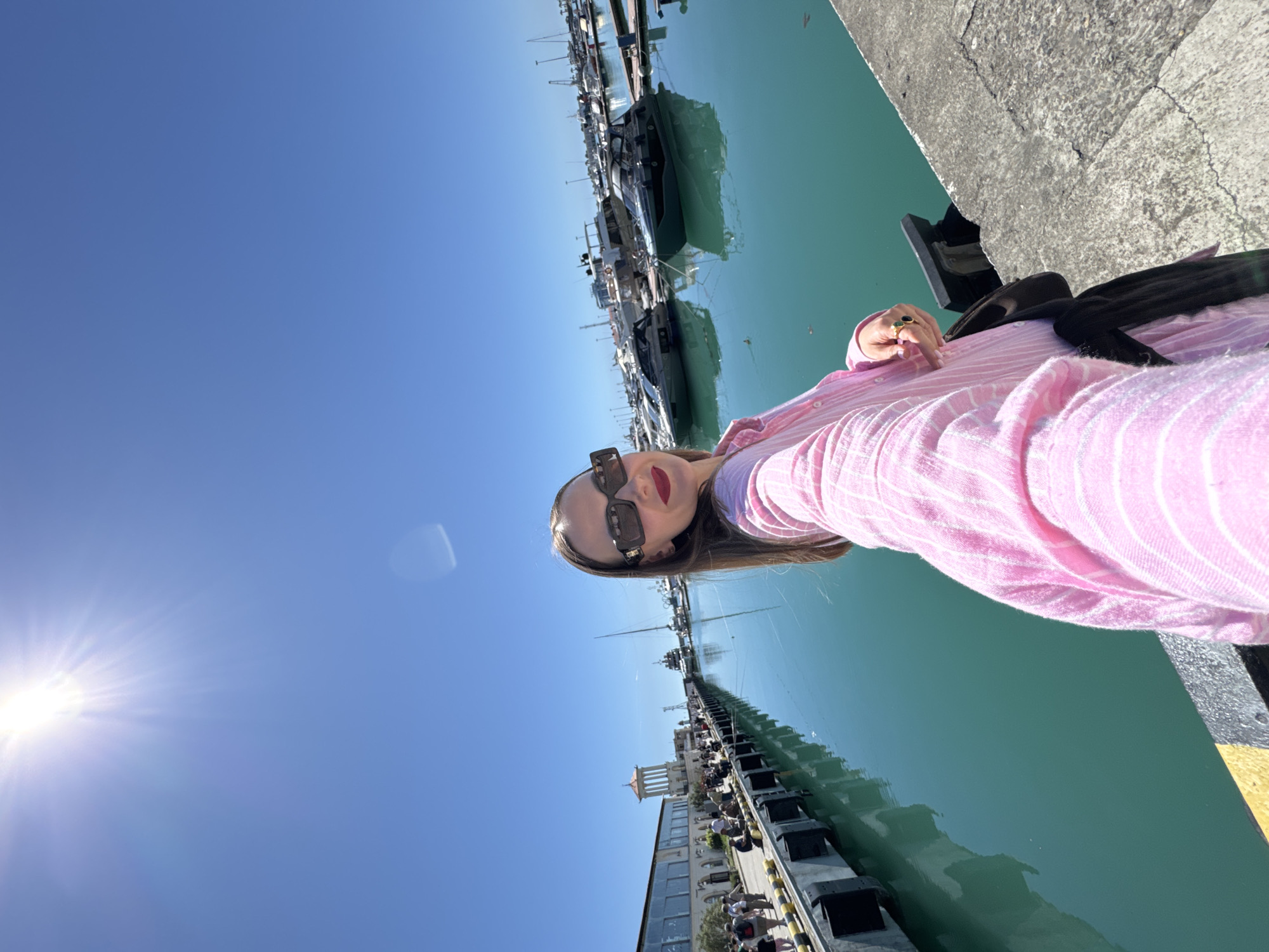 The image is a brightly lit, horizontally rotated selfie of a woman at a marina, likely in Sochi, Russia, on a clear, sunny day. The woman, positioned on the right side of the frame, wears a pink and white striped shirt, dark rectangular sunglasses, and dark red lipstick, looking directly at the camera. Her long brown hair frames her face. On her left hand, a ring with a green stone is visible. A dark strap or garment is seen over her shoulder, resting against her pink shirt.
To her left, the calm, turquoise-green water of the marina reflects the boats docked along the concrete quay. Numerous boats and yachts of varying sizes line the waterfront, extending into the background. Along the far side of the marina, several buildings are visible under the clear blue sky. One prominent structure features a white, classical-style tower or rotunda. People can be seen walking or gathered along the distant promenade.
The sun is bright and high in the sky, creating a strong lens flare in the upper left quadrant of the image. The vibrant colors of the water, sky, and the woman's attire contribute to a cheerful and leisurely atmosphere. There is no visible text in the image.