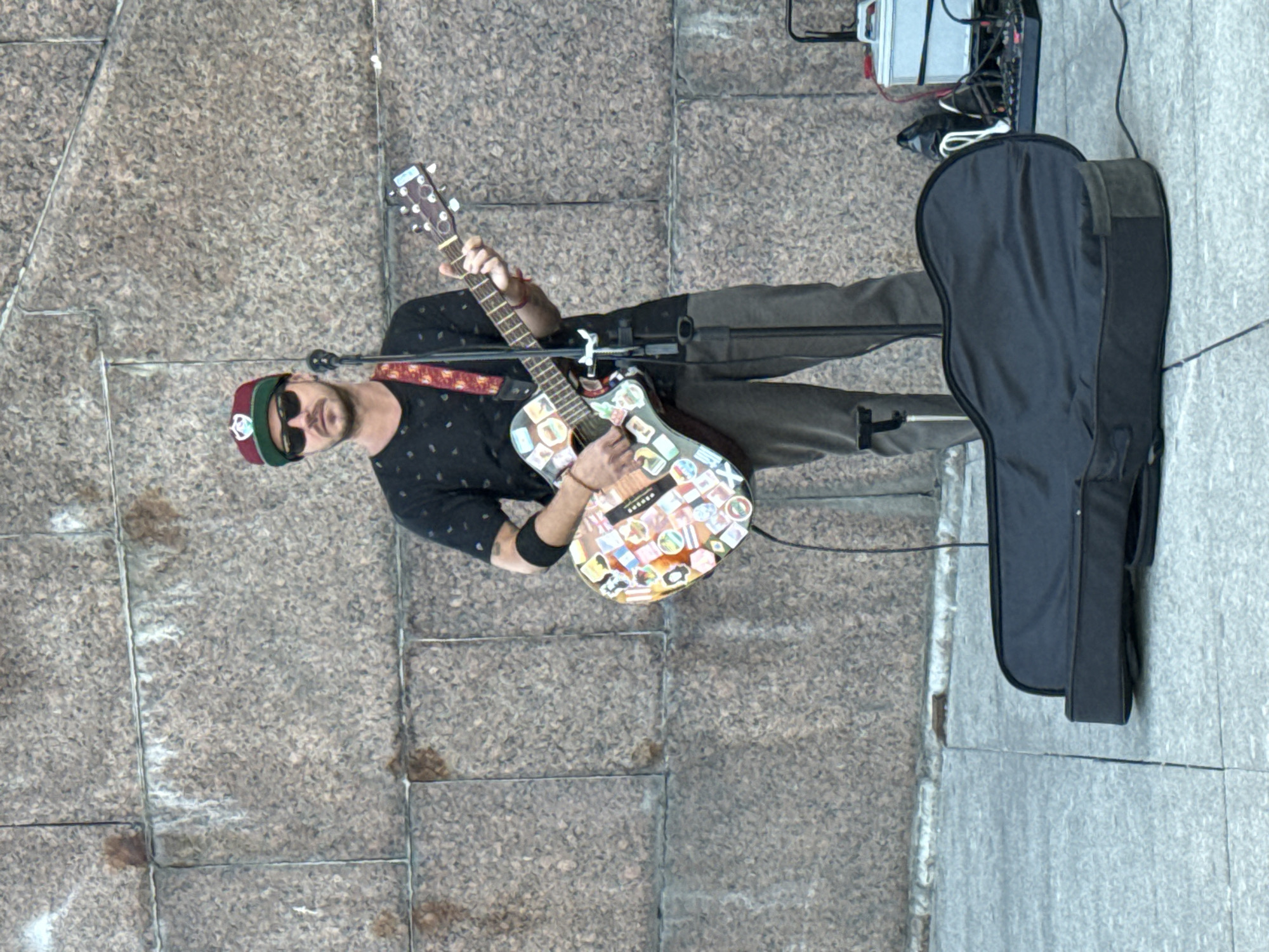 A man stands on a sidewalk, playing an acoustic guitar in front of a large, textured stone wall in Saint Petersburg, Russia. He wears a black t-shirt with a subtle pattern, dark pants, a baseball cap, and sunglasses. The guitar is adorned with numerous colorful stickers. A microphone on a stand is positioned in front of him, and to his right, a black guitar case lies open on the ground next to a small audio mixer and a portable amplifier. The ground appears to be paved with light-colored rectangular stones.