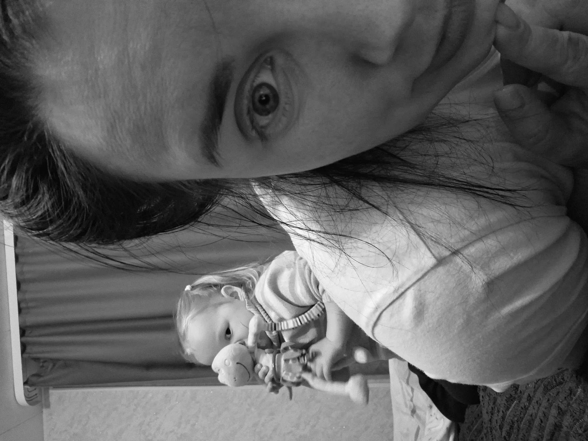 A black and white close-up photograph depicts a woman and a baby indoors. The woman's face, including an eye, is prominently featured at the top of the frame. Her dark hair is visible, and her hand is near her face. The baby is positioned lower in the frame, holding a stuffed animal. The background includes a wall with a patterned surface. The location is in the city of Unknown, Unknown.