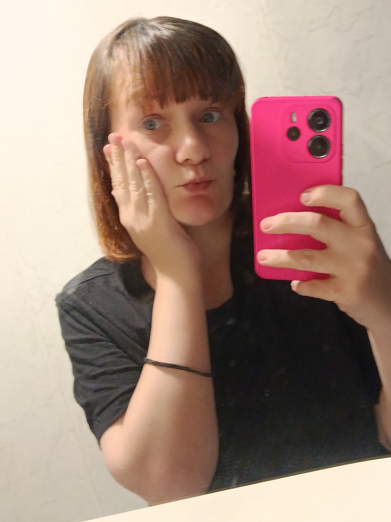 A woman takes a selfie indoors. She has light skin, shoulder-length brown hair with bangs, and is wearing a black t-shirt. Her right hand is touching her face, and she is holding a bright pink smartphone in her left hand. The background is a plain, light-colored wall.