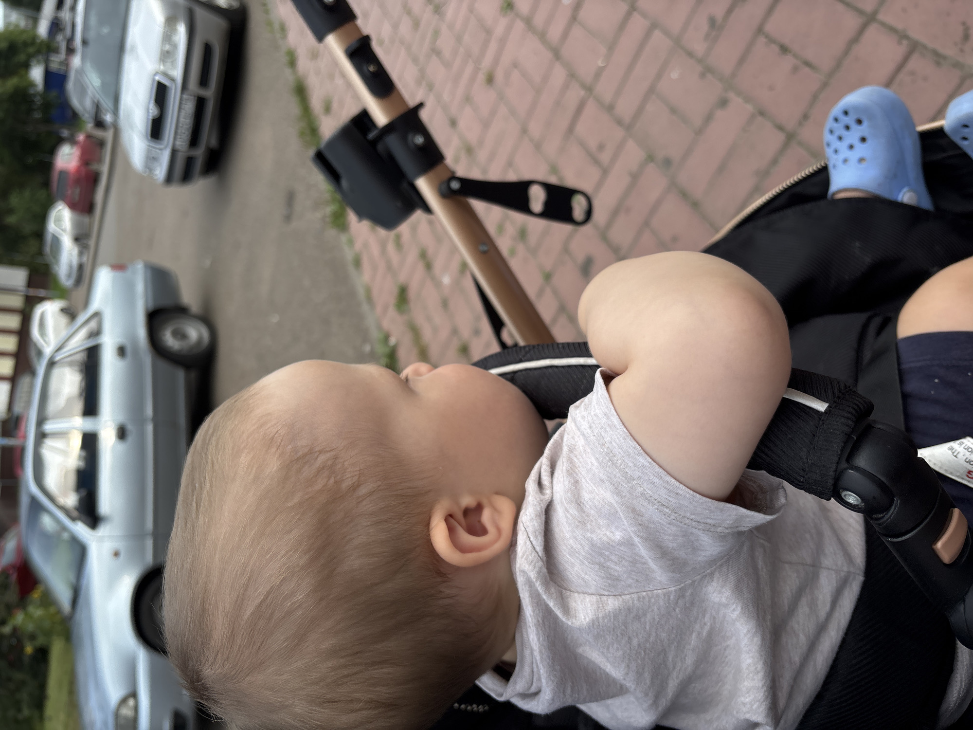 A close-up, eye-level shot shows a young child in a stroller looking to the right. The child has light brown hair and is wearing a light gray t-shirt. The stroller has a black seat and a golden-colored frame. In the background, cars are parked on a street with a brick sidewalk. The scene appears to be in Naberezhnye Chelny, Russia, a city known for its automotive industry and located on the Kama River.