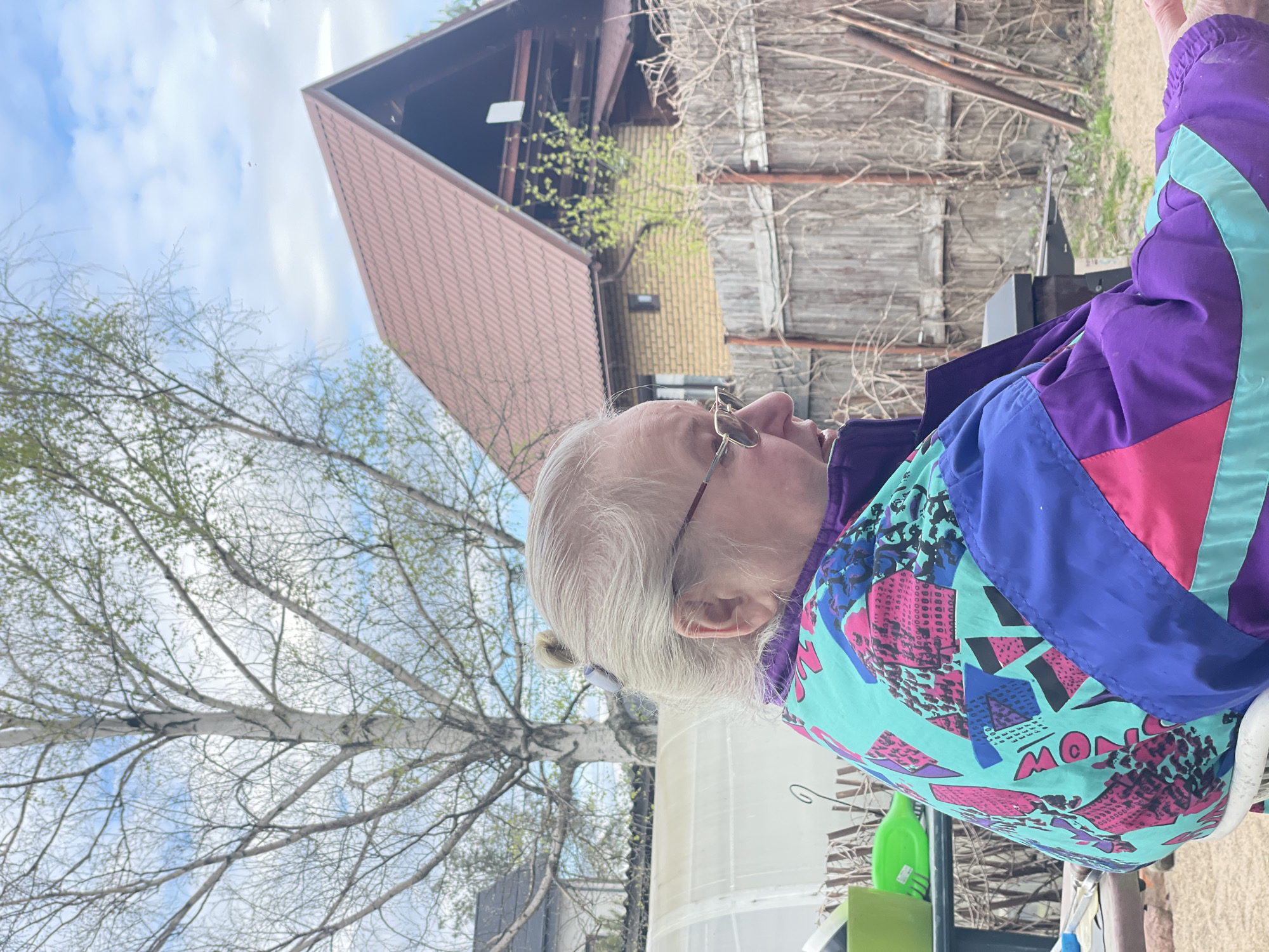 An elderly woman with gray hair and glasses is outdoors, likely in Sestroretsk, Russia, judging by the architecture in the background. She wears a brightly colored jacket. Behind her is a wooden fence, a brick building with a steeply pitched brown roof, and a tree with bare branches. The sky is visible with scattered clouds.