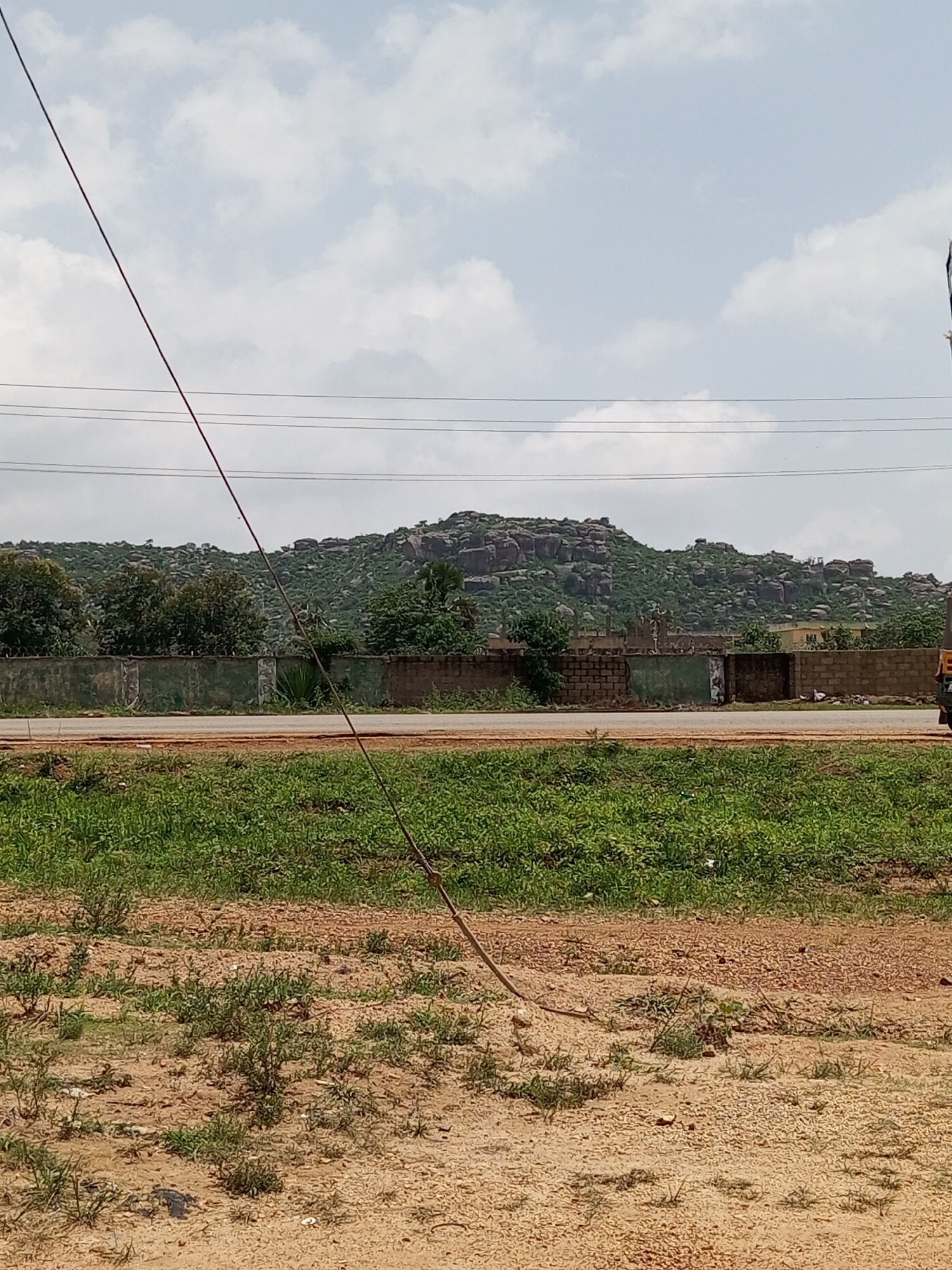The image depicts a landscape scene in Unguwar Saya, Nigeria. The foreground is dominated by dry, sandy earth with sparse patches of green grass and vegetation. A single wire runs diagonally from the top left to the bottom right of the frame, appearing to be a power line or telephone wire.
In the middle ground, a paved road runs horizontally across the image, bordered by a low, greenish wall constructed of concrete and brick. Beyond the wall, a dense area of trees and foliage can be seen, with some buildings partially visible.
The background features a prominent rocky hill or mountain range, covered in green vegetation and dotted with large boulders. The sky above is partly cloudy with a light blue hue, suggesting daytime. The overall impression is one of a rural or semi-rural environment with natural and man-made elements coexisting. No people are visible in the image, and the activity or event taking place is not evident. The weather appears to be fair. No discernible text is present in the image.