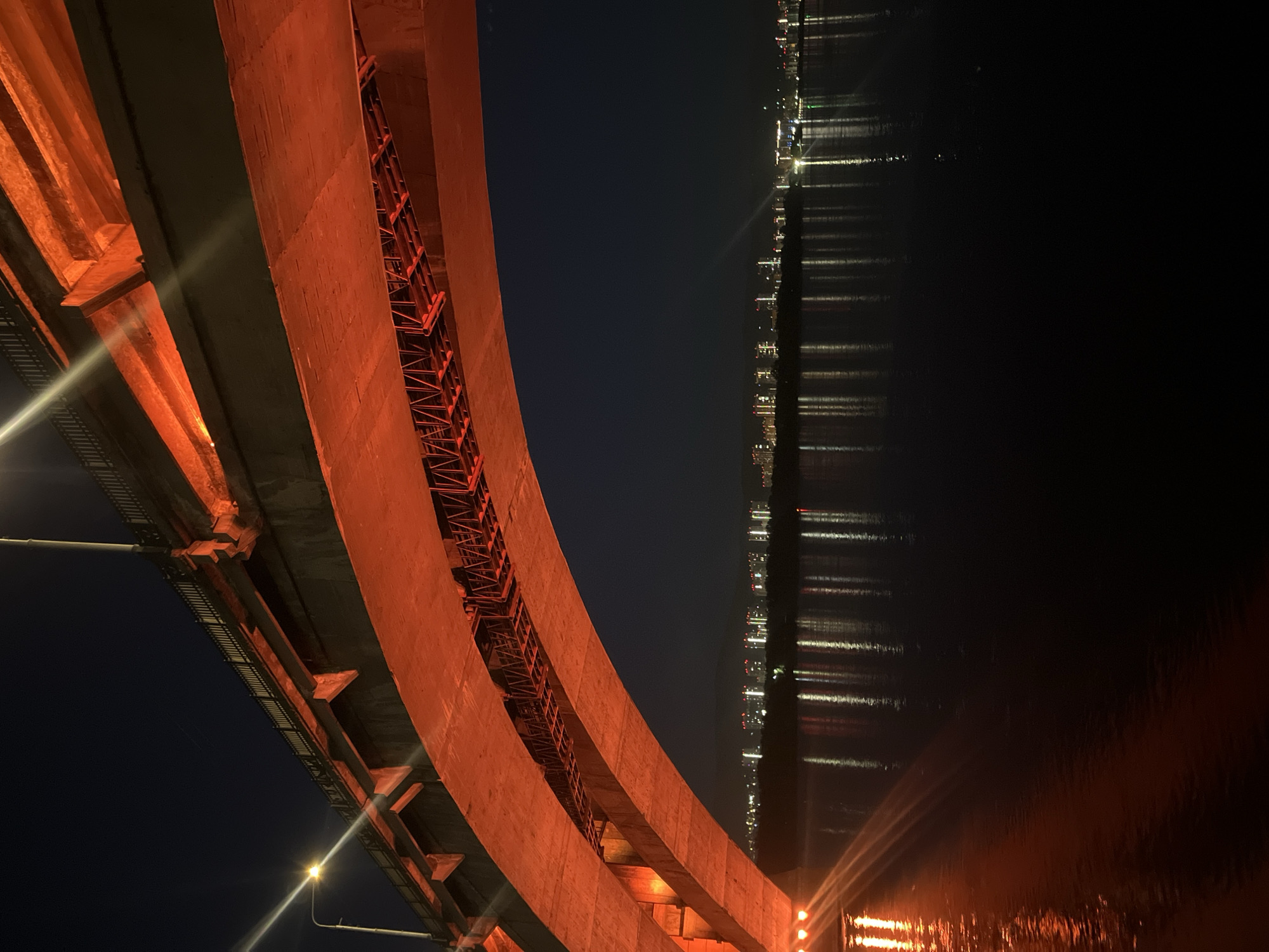 This media file captures a nighttime view from beneath a large, curving bridge structure in an urban setting. The massive concrete bridge, which occupies the left and central portion of the frame, is illuminated with a vibrant orange or reddish light, highlighting its rough texture and the intricate network of support beams and railings beneath its main deck. The perspective is upward-looking, emphasizing the scale and architectural details of the bridge.
To the right, a dark body of water stretches horizontally, reflecting numerous distant city lights or other bridge lights as elongated streaks across its calm surface. The reflections suggest a clear night. The dark night sky is visible above the water and between the bridge structures. Several bright artificial light sources, likely streetlights, are visible in the bottom-left, casting strong rays.
The scene conveys a sense of quiet grandeur, showcasing the blend of engineering and natural elements at night. There are no people visible, and no specific activity or event is taking place. The time of day is clearly night due to the dark sky and reliance on artificial lighting. The location context provided is Krasnoyarsk, Russia. No visible text is present in the image.