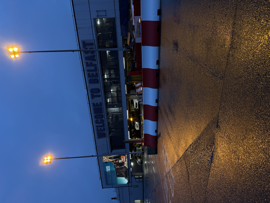 This image displays the exterior of Belfast International Airport, located in Crumlin, United Kingdom, during twilight or night conditions. The sky is dark blue, and the asphalt pavement is wet, reflecting the artificial lights.
The primary subject is the airport terminal building, which features a prominent sign across its facade stating "WELCOME TO BELFAST" in large blue lettering. To the left of this sign, a large billboard displays an advertisement for "BRILLIANT CITY BREAKS AT UNBEATABLE PRICES" by "Jet2 Holidays," alongside an image of an ancient-looking stone archway. The ground level of the building includes a brightly lit, covered drive-through area where at least three light-colored vehicles, including one SUV, are visible. A person wearing a high-visibility vest is partially discernible near the vehicles on the left side of this area.
Two tall, grey metallic light poles, each topped with four amber floodlights, stand in the foreground and midground, casting strong illumination onto the wet pavement. A continuous line of red and white plastic traffic barriers separates the foreground area from the building's immediate entrance. Behind these barriers, a dark blue or black tarpaulin covers a low structure, and a yellow sign with the word "MINSTER" is visible on the right. The wet pavement exhibits reflections of the lights, with faint surface cracks and subtle white markings.