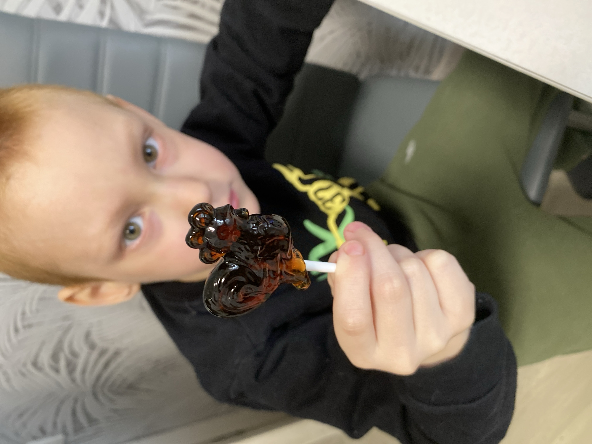 A young, fair-skinned, red-headed boy is holding a dark brown, rooster-shaped lollipop on a white stick in his right hand. The boy wears a black long-sleeved shirt with yellow and green design on the front, and olive-green pants. The background includes a gray padded bench with a white surface partially visible in the upper right corner. The location is Noviy, Russia.