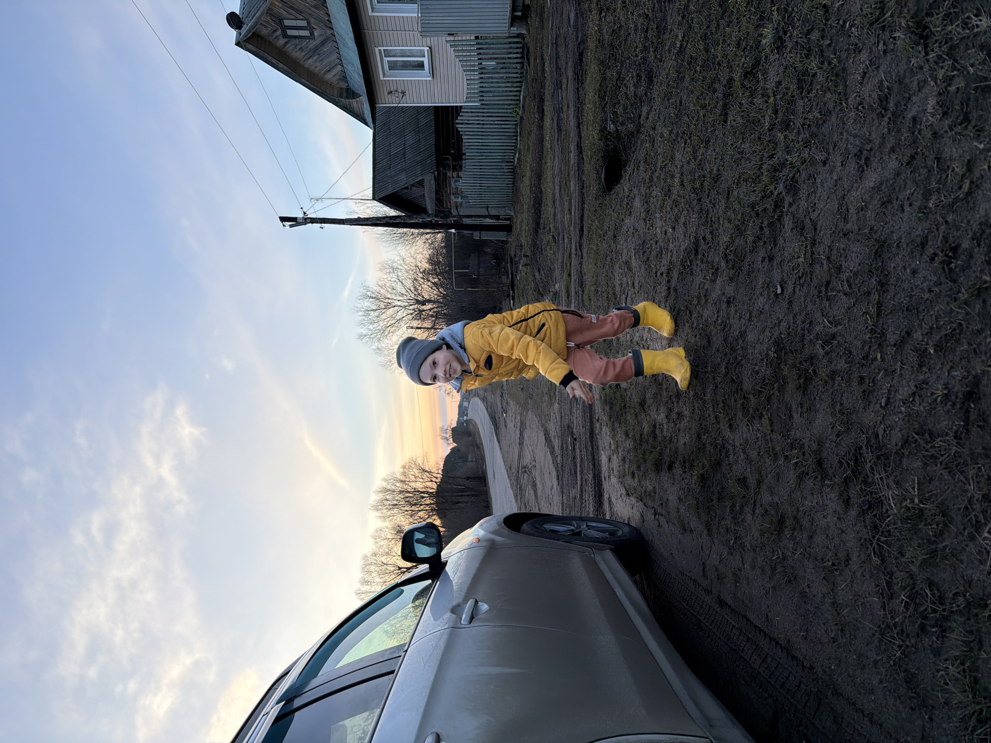 A photograph taken in Дулесово, Russia, shows a young child in a yellow jacket, brown pants, and yellow boots. They also wear a grey beanie. The child is on a dirt road beside a grey car. The background includes a house, a telephone pole, bare trees, and a sky with soft, diffused light.