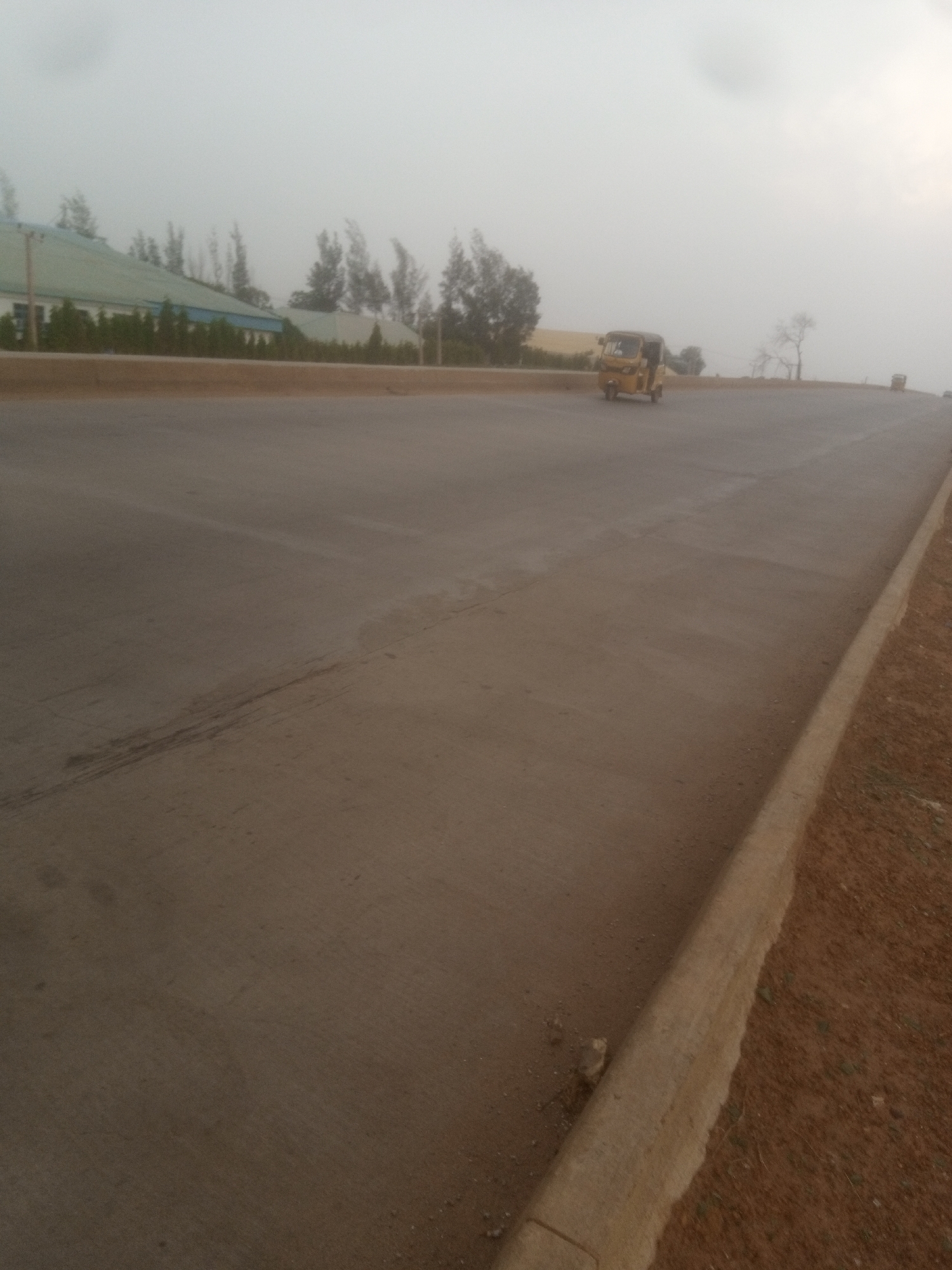 The image depicts a hazy, overcast day in Sabon Gari, Nassarawa, Nigeria. The foreground is dominated by a wide, paved road, likely a highway or major thoroughfare, with a distinct curb on the right side separating the road from a reddish-brown dirt embankment. The road surface appears relatively new, with some tire marks visible. In the mid-ground, a yellow three-wheeled vehicle, possibly a tuk-tuk or auto-rickshaw, is traveling on the road. Further in the background, buildings with green roofs are visible, along with sparse vegetation and trees. The overall atmosphere is subdued due to the overcast conditions.