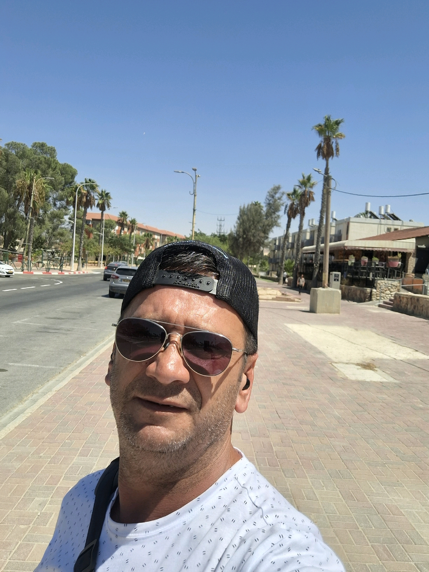 A man wearing sunglasses and a baseball cap takes a selfie on a sunny day in Yerocham, Israel. He is standing on a pedestrian walkway paved with light brown and tan rectangular tiles. To his left, a street with two lanes of traffic runs parallel to the walkway, bordered by a red and white striped curb. Several cars are visible on the street, with a silver sedan in the foreground and a white car further ahead. On the right side of the street, there are buildings with awnings, trees, and a tall utility pole. Palm trees are scattered throughout the background. The sky is clear and blue. The man is wearing a white t-shirt with a subtle dotted pattern and a dark strap from a bag over his shoulder. He has an earpiece in his right ear.