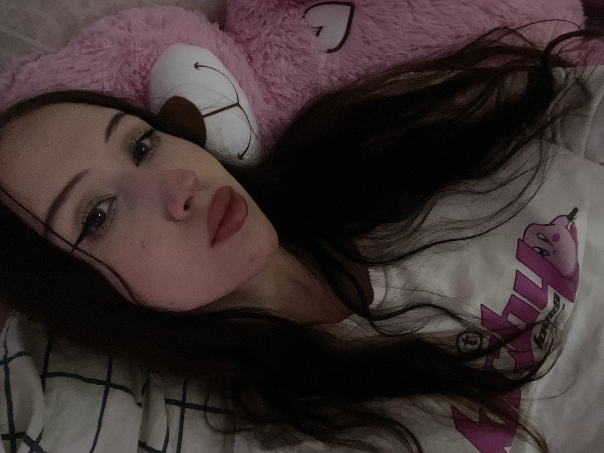 The image is a selfie taken by a young woman with long, dark hair. She is lying down, with a pink teddy bear and a plaid blanket visible in the background. The woman is wearing a white t-shirt with a graphic print that appears to feature a cartoon character. The lighting is dim, suggesting the photo was taken indoors, possibly in a bedroom. There is no indication of the specific location within Votkinsk, Russia.