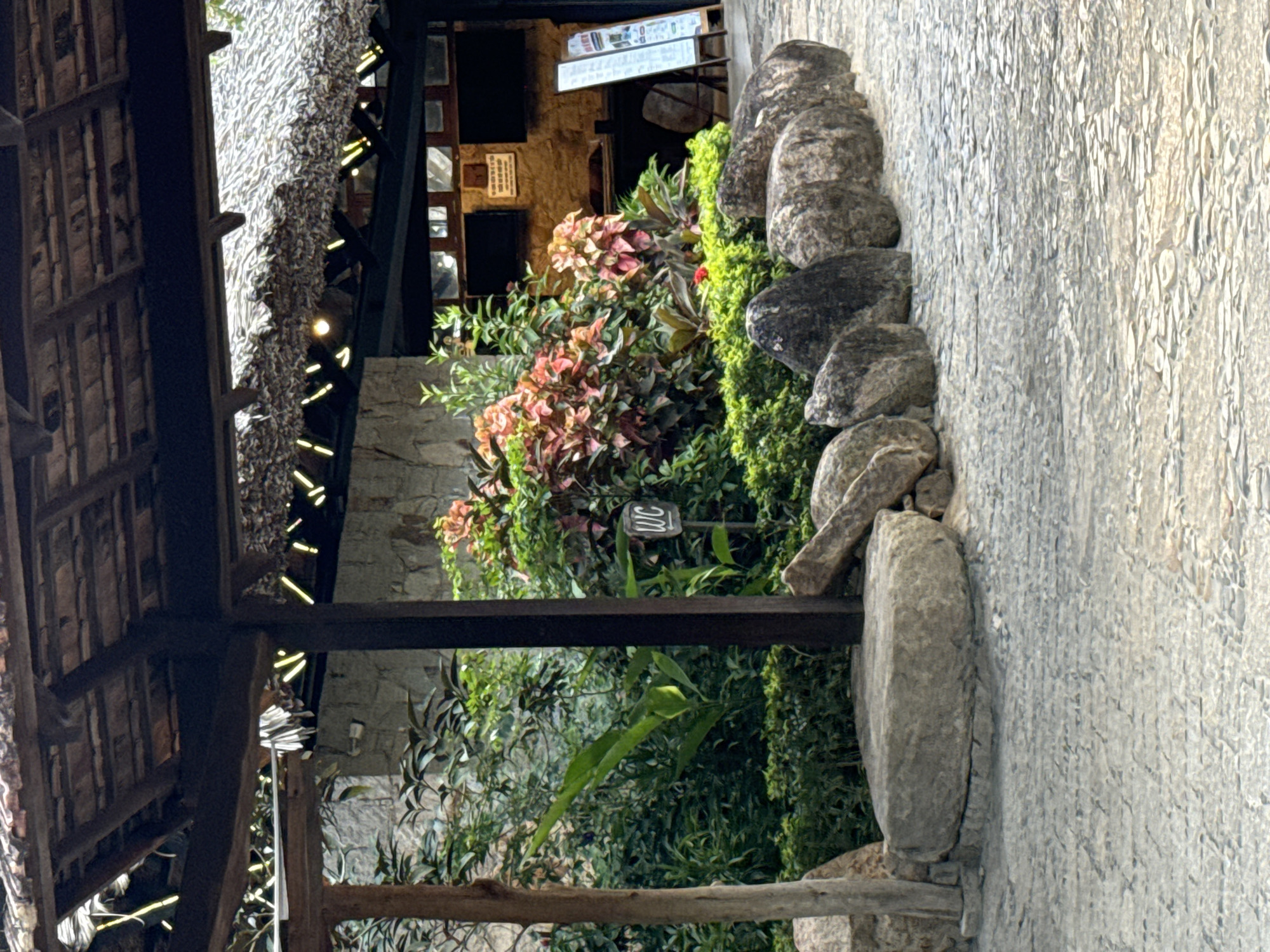 This outdoor shot from Tay Nha Trang Ward, Vietnam, shows a thatch-roofed structure at the left, partially framing the scene. Next to this, a wooden post is visible, supporting the roof. Lush greenery fills the space between the roof and a stone wall on the right. A small, black sign with white lettering, possibly indicating a toilet, is nestled within the greenery. A stack of grey stones, placed vertically, borders the wall. The wall itself is textured and light grey.