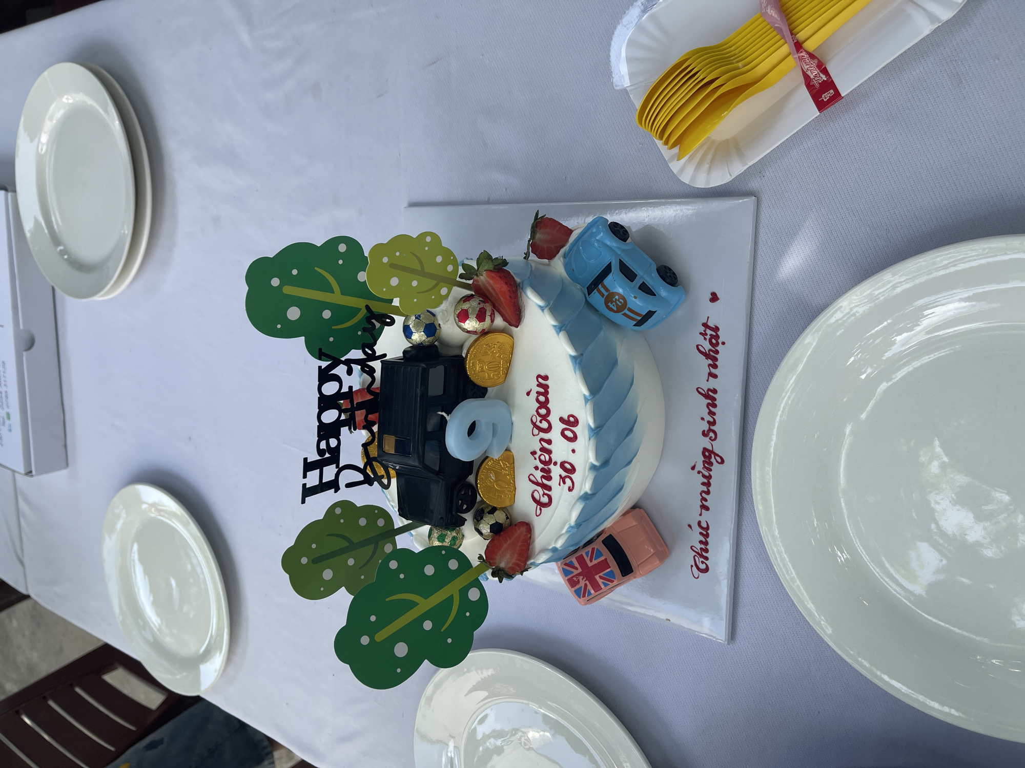 A close-up, eye-level shot shows a birthday cake topped with a toy truck, trees, soccer balls, strawberries, gold coins, and candles. A blue "9" candle sits prominently on the cake, which is decorated with blue and white frosting and red lettering that reads "Chien Toan 30.06" and "Chúc mừng sinh nhật". Two toy cars, one pink with a British flag design and another blue, are placed on the cake's base. The cake sits on a white platter, which is on a white tablecloth. In the background, several white plates are stacked, and a cardboard box is visible. To the right of the cake, a white paper plate holds a bundle of yellow plastic cutlery tied with a pink ribbon. The overall scene appears to be set at an outdoor birthday celebration, possibly in a residential area of Phu Thuy Ward, Vietnam, given the Vietnamese text on the cake.