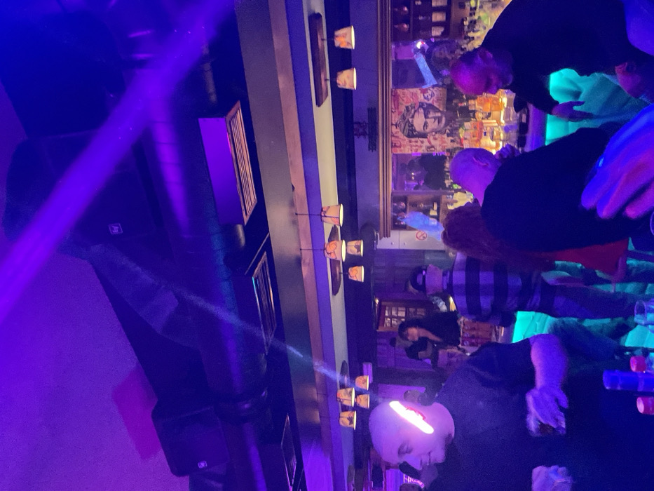 The image captures an indoor nighttime scene, likely within a bar or club in Drapetsona, Greece. The photograph is oriented vertically, rotated 90 degrees counter-clockwise, with the bar running horizontally across the mid-ground.
The space is illuminated by vibrant purple and blue lighting, creating a dynamic and somewhat dim atmosphere. In the foreground, a bald man holds a glass with a drink and rests his hand on the bar. A distinct, elongated white light reflects prominently across his head. Next to him, another person is partially visible, also at the bar.
Behind them, several other patrons are present. A man wearing a plaid shirt and a cap stands further back, and another person with reddish hair is partially obscured. Towards the upper right of the frame, another individual leans over the bar.
The bar counter features drinks and bottles. Above the bar, a series of small, cup-like decorative lights provide a warm glow. A large, framed artwork or poster with a stylized portrait in a pop-art style is visible on the wall behind the bar. A clear white "No smoking" sign is also visible on this wall.
On the left side of the image, appearing to be part of the ceiling or upper wall structure, black speakers (one with a faint "EV" logo) and air ducts are present. A prominent beam of purple light cuts across this area, adding to the club-like ambiance. The scene depicts a social gathering in an energetic, artificially lit environment.