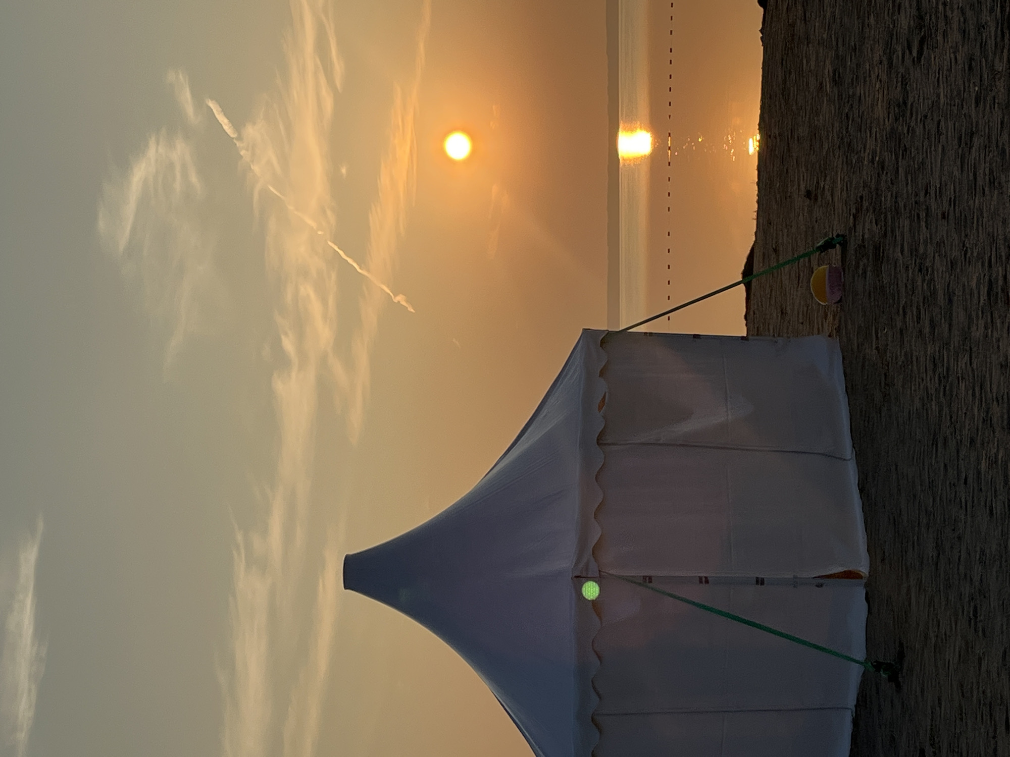 A white tent with a conical roof is pitched on a sandy beach at sunset. The tent is supported by green poles. A small pink and yellow bucket sits in the sand near the tent. The sun is setting over a calm body of water, casting a golden reflection. The sky is orange and yellow with some wispy clouds. A contrail from an aircraft is visible in the sky. The image was taken in Surakovo, Russia.