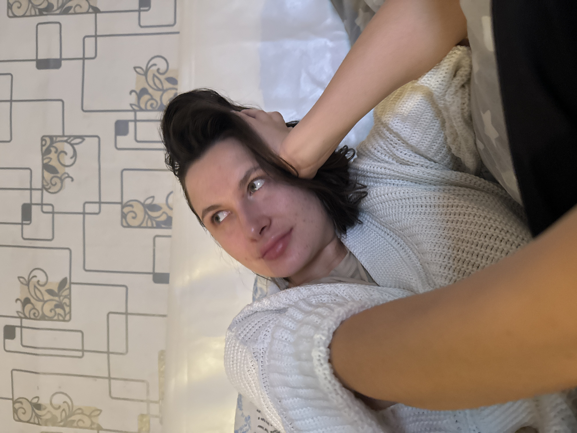 A woman with dark hair, wearing a white cardigan, is sitting and looking to the left of the frame. Her right hand is on her head. The background consists of a wall with a patterned wallpaper and a white sheet. The city and specific location are unknown.