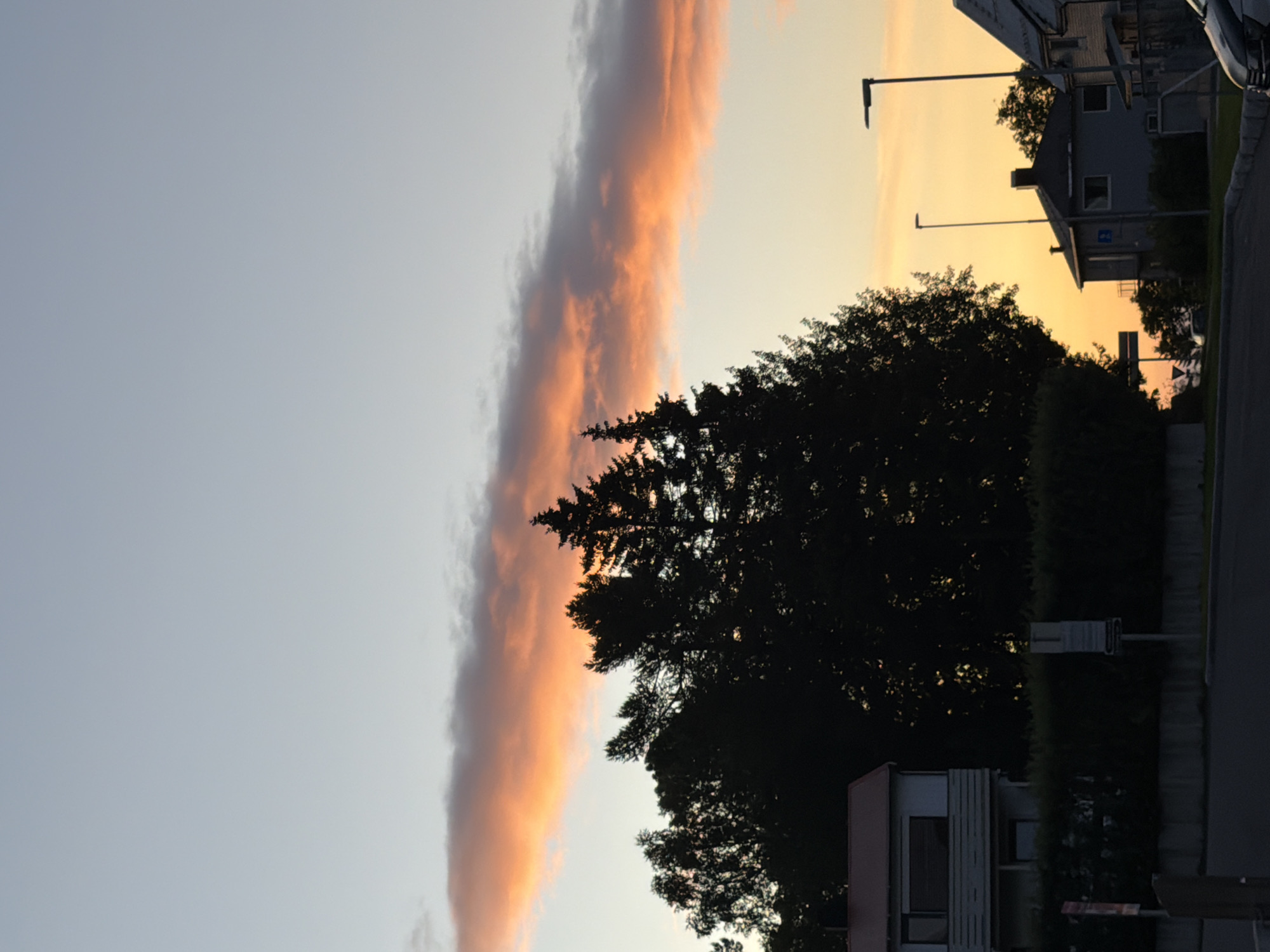 In Harstad, Norway, a street scene unfolds during a twilight period. The sky is a gradient of soft blue at the top, transitioning to a warm orange glow near the horizon, illuminated by a long, thin cloud band tinged with pink and orange. Silhouetted against the sky are tall evergreen trees, their dark forms contrasting with the vibrant clouds.
In the foreground, a residential area is visible. A light-colored house with a red roof is situated to the left, featuring a multi-story structure with a prominent window on the upper level. Adjacent to it, a well-maintained hedge forms a dark green barrier. Further to the right, other houses with pitched roofs are partially visible, their details softened by the low light. Streetlights stand along the road, their poles reaching upwards. The road itself is paved, with a curb marking its edge. The overall atmosphere is one of quiet dusk in a suburban setting.