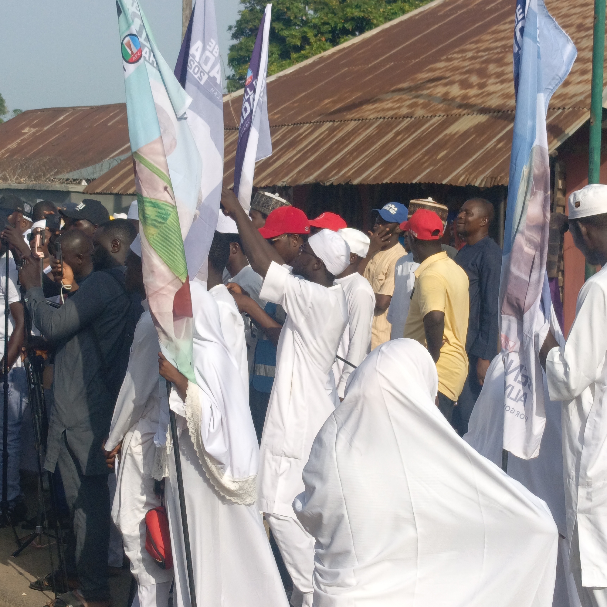 In Akwanga, Nigeria, a crowd of people are gathered outdoors. Many are wearing white clothing, including hooded robes. Some wear red or blue caps. Several people hold up flags with political logos and names. Others are taking pictures with their phones. A building with a corrugated metal roof is in the background.