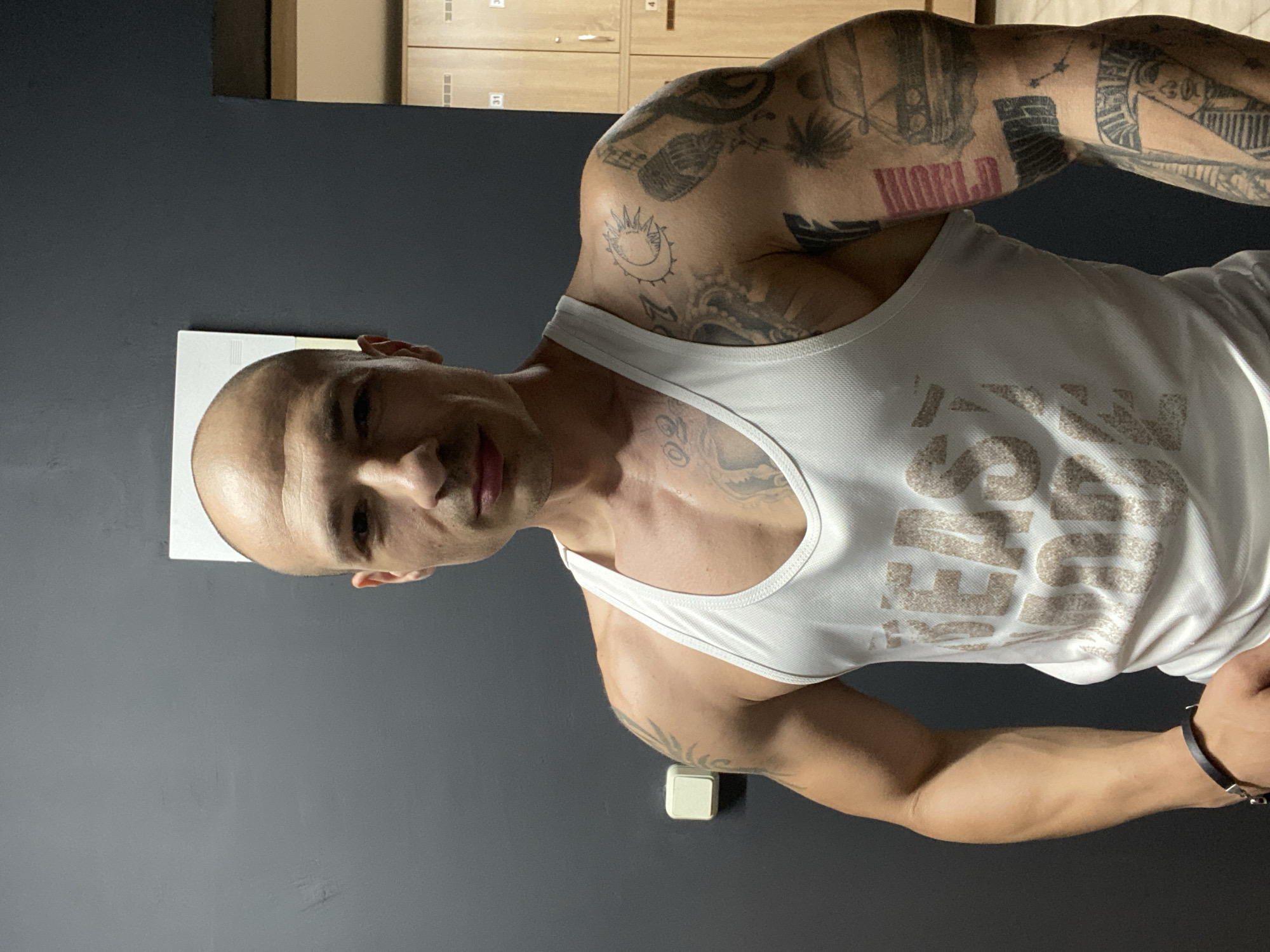 A man with a shaved head and a muscular physique is captured indoors, likely in a gym or changing room in Pleven, Bulgaria. He is looking directly at the camera with a confident expression and a slight smile. He wears a white sleeveless tank top, which has a faded, indistinct light brown graphic print on the chest.
Both of his arms are heavily adorned with black ink tattoos, depicting various symbols and patterns. On his left bicep, the word "WORLD" is distinctly tattooed in red or pink letters. A sun and crescent moon design is visible on his right shoulder, among other intricate patterns. A dark bracelet with metallic accents is on his right wrist.
The background features a plain dark gray wall. In the upper right corner, several light wooden lockers with numbers (e.g., '31' and '4') are partially visible, reinforcing the indoor setting of a changing room or athletic facility. A white rectangular object is mounted on the wall to the left of the man's head, and a smaller white square object is positioned on the wall below his right arm. The scene is illuminated by indoor lighting, casting soft shadows. The man appears to be posing for the photograph.