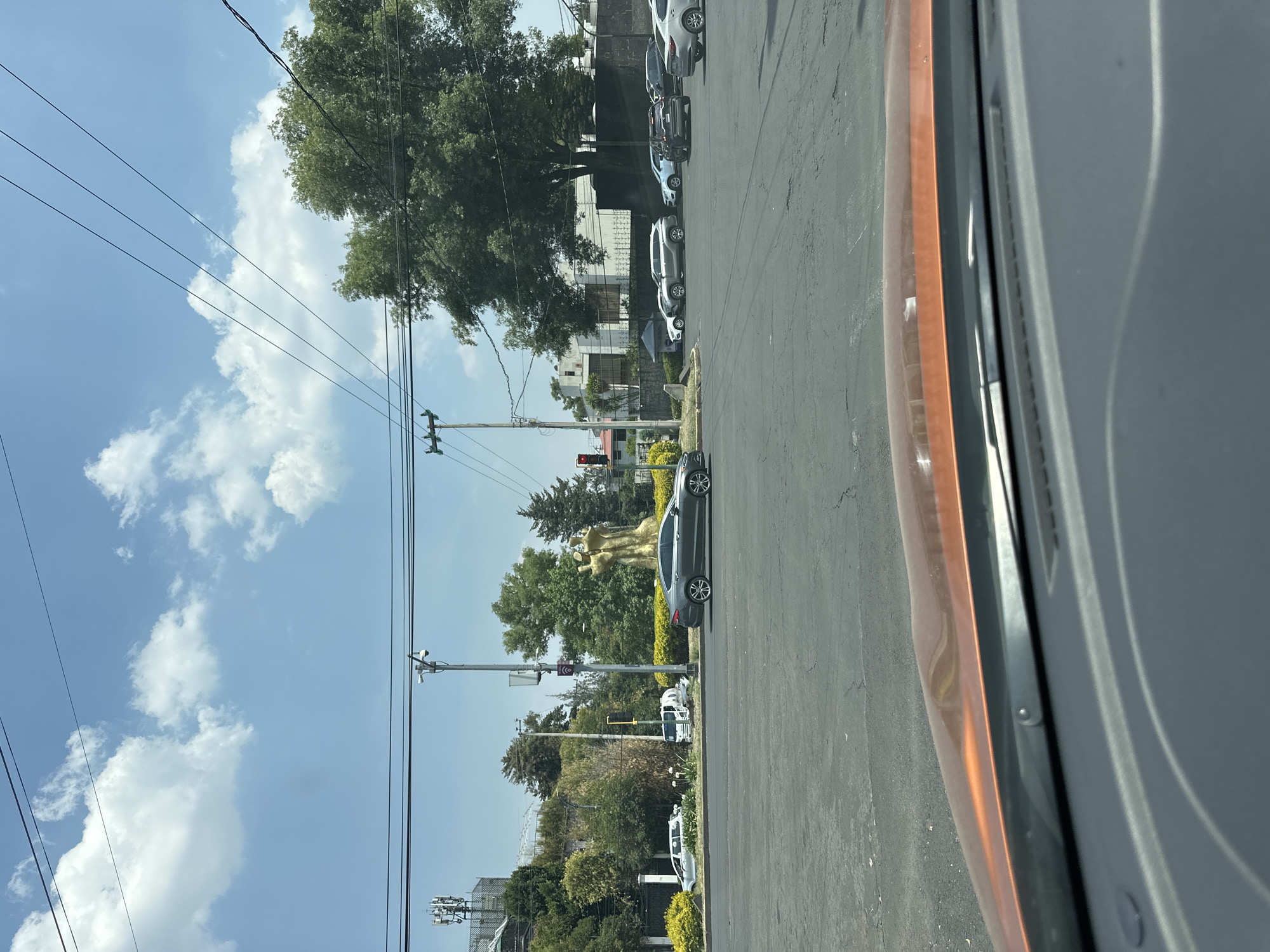 The image displays a daytime street scene, likely from the perspective of someone inside a vehicle, given the visible orange-brown car trim in the foreground on the right. The sky, occupying the upper portion of the frame, is a clear blue with scattered white, fluffy clouds, indicating good weather.
Several power lines and utility poles crisscross the sky. Below, a paved road stretches across the middle of the frame. A line of cars, mostly light-colored sedans and SUVs, are parked along the right side of the road. Beyond the parked vehicles, a dense array of green trees and foliage is prominent, partially obscuring some buildings in the background. A notable feature is a large, golden, abstract sculpture, possibly depicting two figures, standing amidst the trees and a low hedge or fence. Further down the street, a traffic light is visible on a utility pole. On the far right, parts of a chain-link fence and telecommunications equipment on a pole can be seen.
The scene suggests an urban or suburban setting in Mexico City, Mexico, under bright, sunny conditions. No people or visible text are clearly discernible.