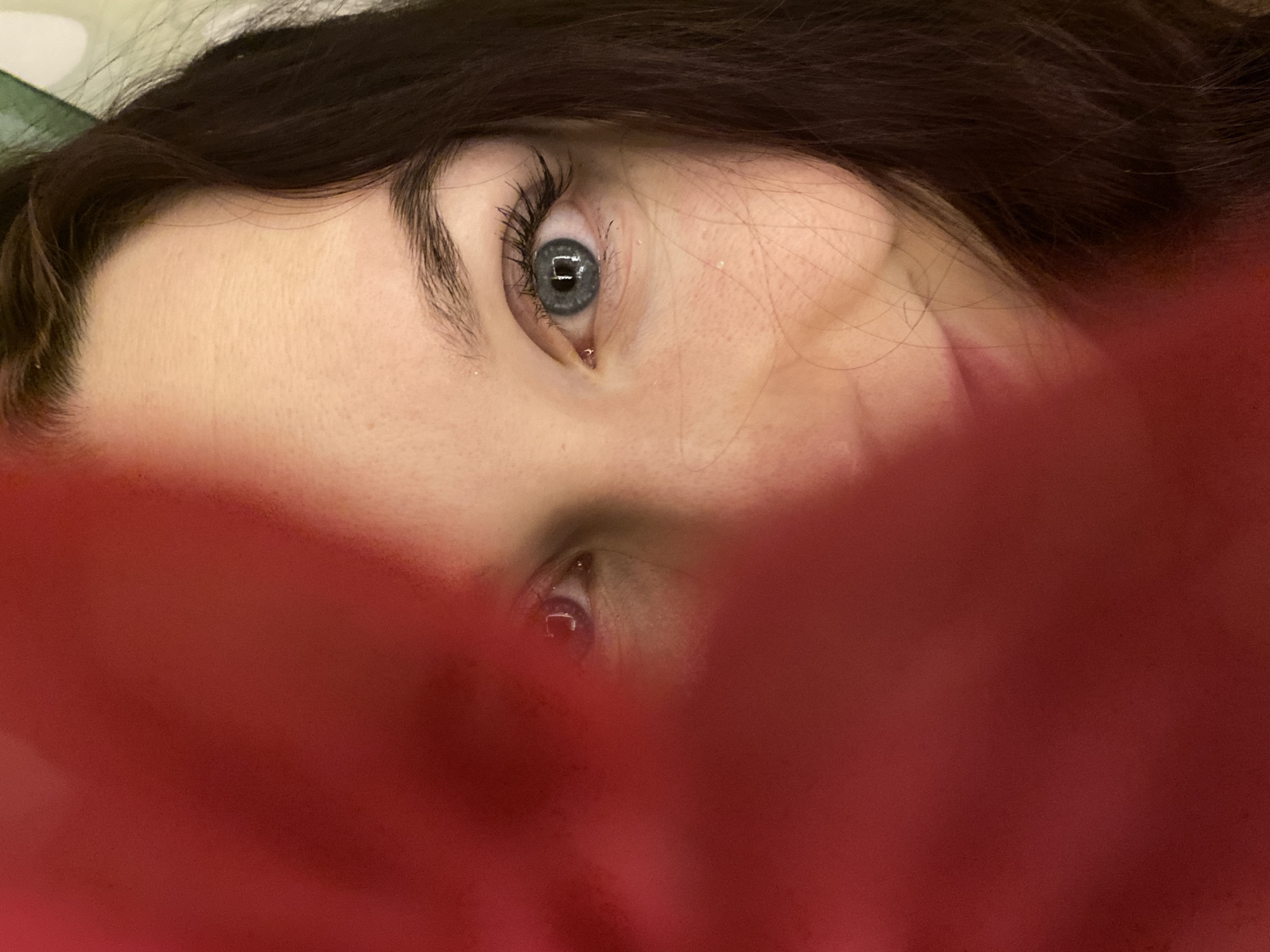 A close-up, high-angle shot captures a person's face, possibly taken in Chaikovsky, Russia. The individual, appearing to be female, has dark brown hair that frames the upper part of her face. Her fair skin is visible around her eyes and forehead. The most prominent features are her bright blue eyes, which are wide open and looking directly or slightly upwards towards the viewer. The eyes are framed by dark, defined eyebrows and long, dark eyelashes, suggesting mascara.
A large portion of the lower face and the right side of the image is obscured by a vibrant, blurred red fabric or material. This red element dominates the foreground and extends across the bottom of the frame, creating a sense of being partially hidden or covered. Only the upper half of the face, specifically the forehead, eyebrows, and eyes, is clearly visible. The reflection of a rectangular light source is visible in both pupils.
The background is mostly dark and out of focus, with a subtle hint of a lighter, possibly green-tinged surface in the upper left corner, suggesting an indoor setting. The overall mood is intimate and focused on the eyes. No specific activity other than looking is depicted, and the expression is neutral. There is no visible text in the image.