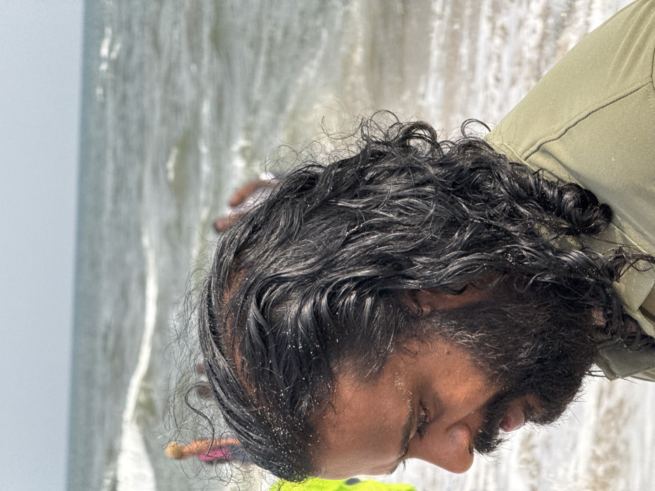 The image presents a close-up, side-profile view of an adult male at a beach location specified as Mopo Onijebu, Nigeria. The man, positioned in the foreground and facing left, has dark, long, wet, curly hair and a full dark beard. Both his hair and forehead display visible specks of sand. He is wearing a light olive green collared garment, with his upper torso partially visible. His gaze is directed downwards. In the blurred background, a large body of water, indicative of an ocean, features breaking waves and foam under a light, clear sky. Several indistinct human figures are discernible in the water and near the shoreline in the mid-ground, suggesting the presence of other individuals at the beach. The scene is illuminated by bright daylight, consistent with an outdoor coastal environment.
