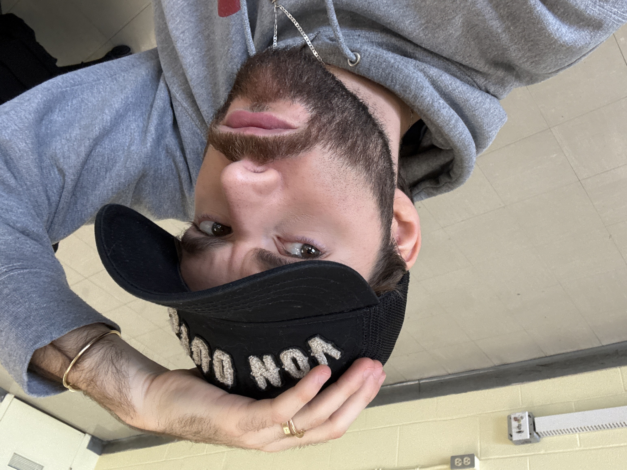 A selfie of a man with a beard and mustache wearing a grey hooded sweatshirt and a black baseball cap with white lettering, is positioned upside down in the frame. The man has fair skin and is wearing a gold bracelet on his left wrist, and two gold rings on his left hand. He is holding the cap. A silver necklace hangs from his neck. The background is a light yellow tiled wall with a light fixture and electrical outlet. There is no indication of the location being in Montreal, Canada.