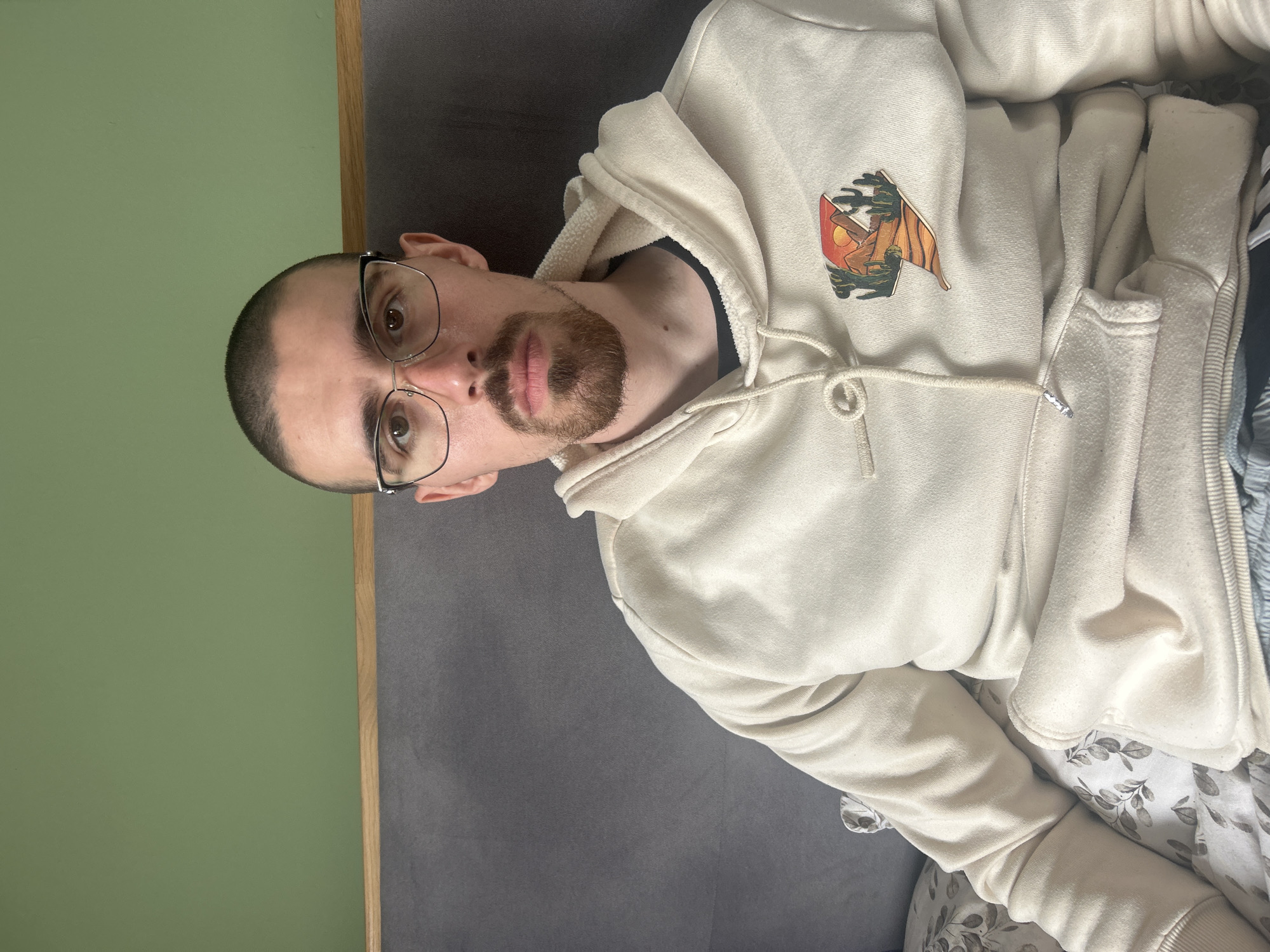A man is seen from a slightly upward angle, wearing glasses and a cream-colored hoodie with a design on the upper left side. He has short hair and a beard. The background consists of a light green wall and a gray surface. The location and city of Burgas, Bulgaria are not determinable from the provided image.