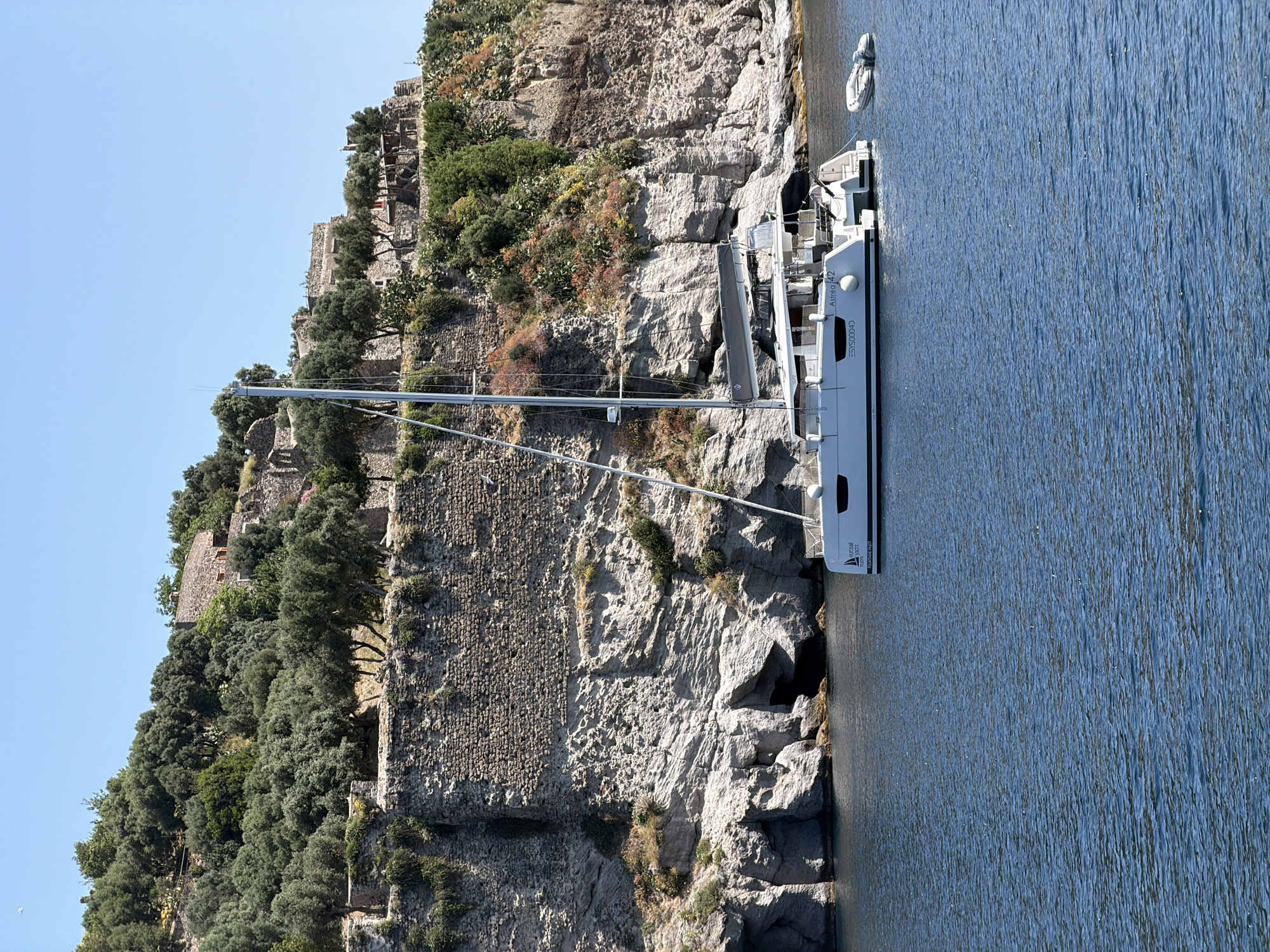The image presents a sideways view of a coastline scene likely in Ischia, Italy, showing a large catamaran-style yacht moored adjacent to a steep, rocky coastline. The coastline is dominated by a high, rugged rock face, part of which appears to be a historic wall or structure with embedded stone and possibly remnants of fortifications. The rock face is partially covered with vegetation, including trees and scrub bushes.
The yacht is a modern design with a tall mast. The yacht is labeled as "Altea 42" with the word "FOUR SEAS YACHT" visible on its hull. A small tender boat is docked just behind the yacht.
The water surrounding the yacht appears calm. The sky is clear and light blue.