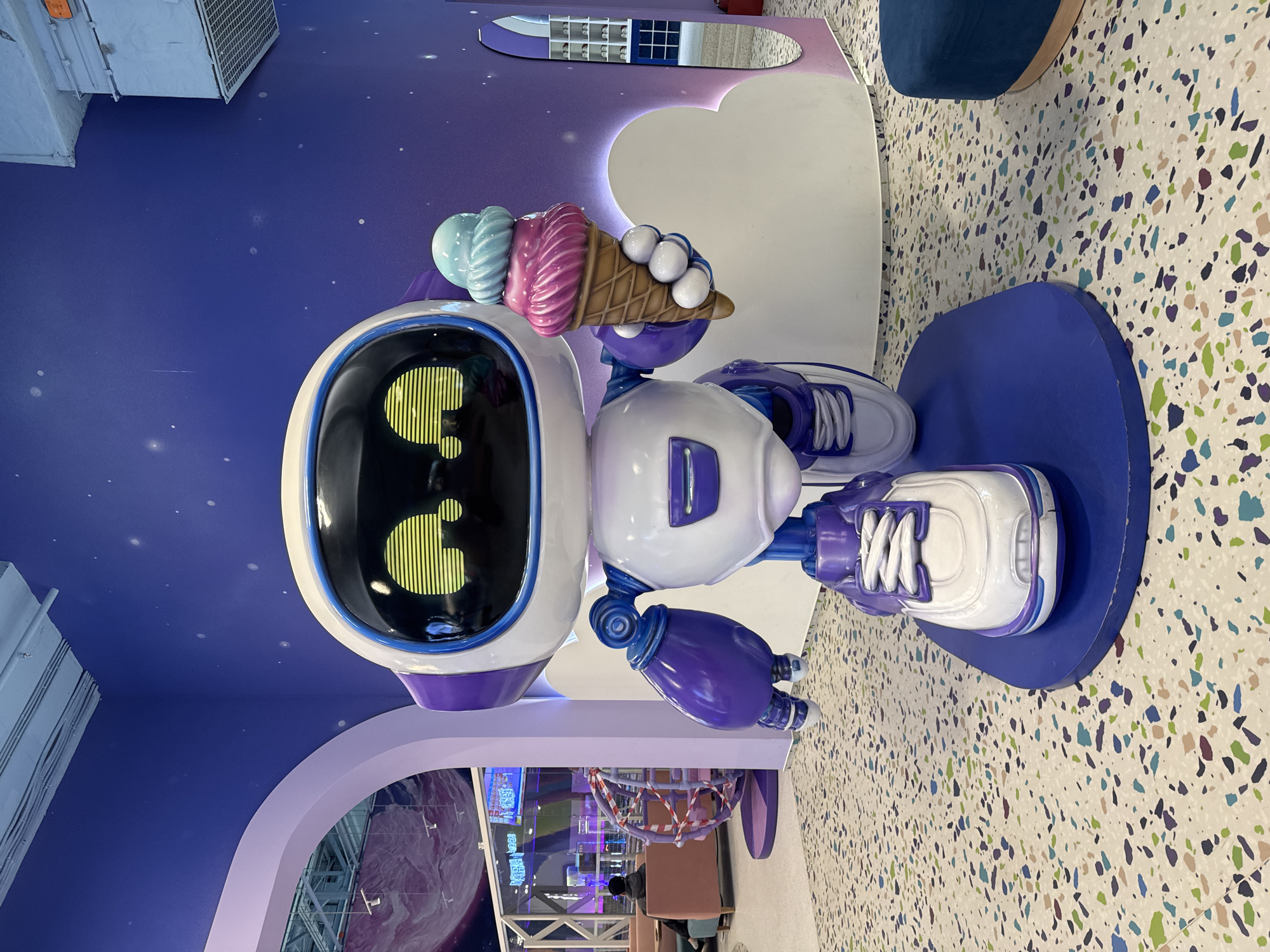 A large, stylized robot sculpture dominates the center of a brightly lit indoor scene, likely a commercial or entertainment space in Saint Petersburg, Russia. The robot, primarily white with purple and blue accents, has a sleek, rounded design. Its head features a black screen displaying a vibrant yellow-green question mark, formed by vertical stripes. In its right hand, the robot holds a large, colorful ice cream cone with three scoops: light blue, pink, and white spheres atop a brown waffle cone. The robot's feet resemble white sneakers with purple laces and soles, standing on a circular blue base.
The background features a dark blue wall speckled with small white dots, resembling a starry night sky. White, stylized cloud shapes are affixed to this wall. To the left, an industrial-style air vent is visible on the ceiling. On the right, the floor is a light-colored terrazzo with scattered chips of blue, green, purple, brown, and beige. A segment of a blue upholstered seat is visible in the upper right.
In the lower left, a light purple archway leads to another area where a large spherical object, resembling a purple planet, is visible. Further back, beyond the archway, reflections of an interior space with bright lighting and structures wrapped in orange and white striped tape can be seen. A person in dark clothing is seated in the background, partially obscured by the robot's base. The overall setting is whimsical and modern, suggesting a fun or child-friendly environment.