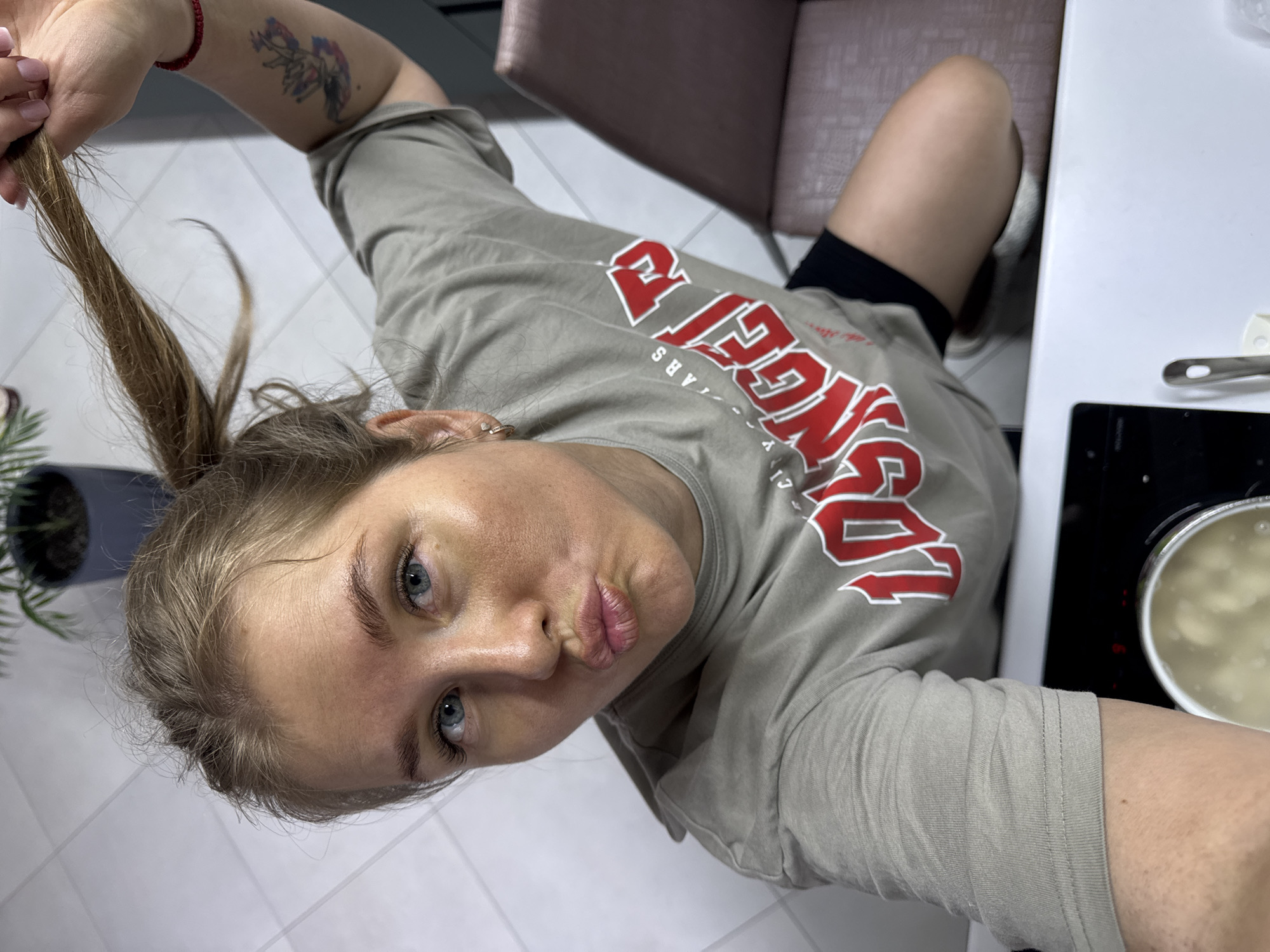 A young woman is featured in a casual selfie, holding a section of her light brown hair with her left hand while making a playful "pouty" expression towards the camera. She has blue eyes and her left arm displays a small, colorful tattoo, along with a red bracelet on her wrist. Her fingernails are painted a light pink. She is dressed in a light olive-green t-shirt with prominent red and white text, which clearly reads "LOS ANGELES" across the chest, with smaller words like "ARS" and "CITY" partially visible below. She is also wearing black shorts.
The scene is an indoor setting, likely a kitchen or dining area in Ферма, Russia, characterized by a light-tiled floor. To her right, a white tabletop holds a metal utensil. A black stovetop is visible, with a pot containing light-colored liquid and food items (possibly dumplings or pasta) boiling on one burner. A portion of a reddish-brown upholstered chair is seen in the background, along with a potted plant on the far left. The natural lighting suggests the photo was taken during the daytime. The overall ambiance is relaxed and lighthearted.