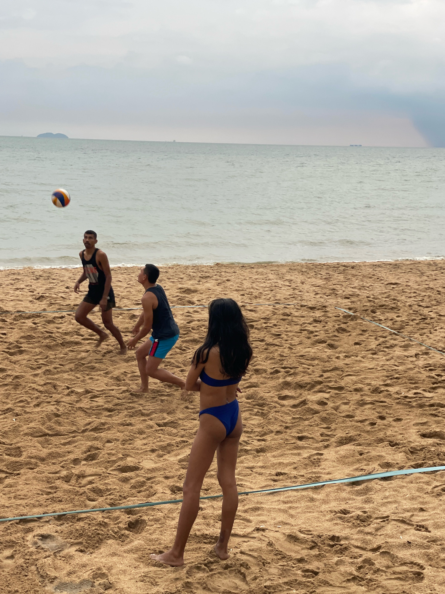 Four individuals are playing beach volleyball on a sandy beach in Pattaya City, Thailand. The sky is overcast, and the sea is visible in the background with a few distant boats and a small island. A volleyball is in mid-air, tossed upwards, with one person appearing to be preparing to hit it. The other three individuals are positioned on the sand, seemingly in a game. The players are dressed in swimwear, with two males wearing tank tops and shorts, and one female wearing a blue bikini. A thin blue line, likely a volleyball net boundary, is visible stretched across the sand. The sand is textured with footprints and divots.