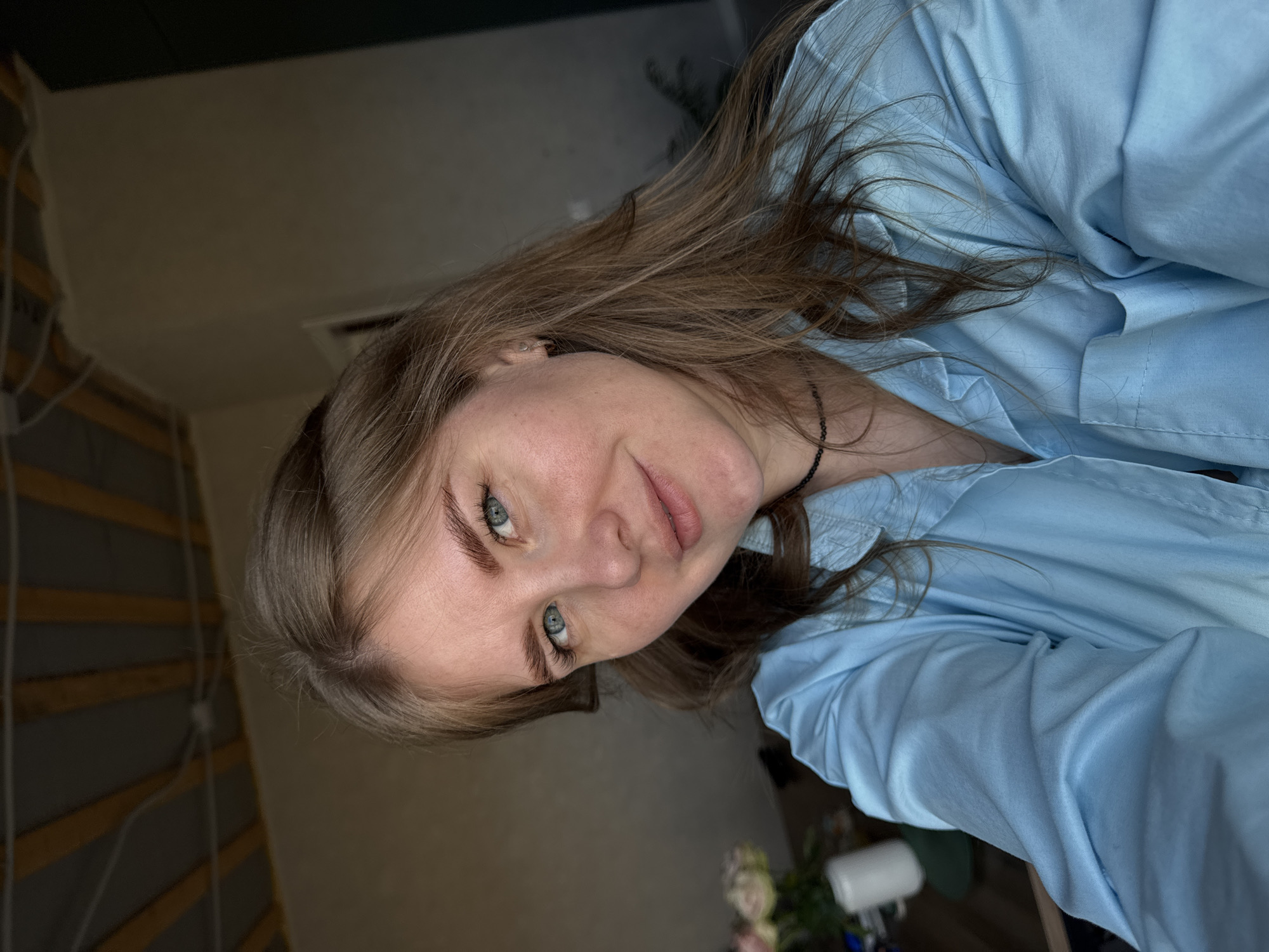 A woman is posing for a selfie in an indoor location. She has light brown hair and blue eyes and is wearing a light blue shirt. The location appears to be a home, with elements of the interior visible in the background. The image's exact location in Gornyi, Russia, cannot be determined from the visual data. There are no apparent actions or interactions besides the woman posing.