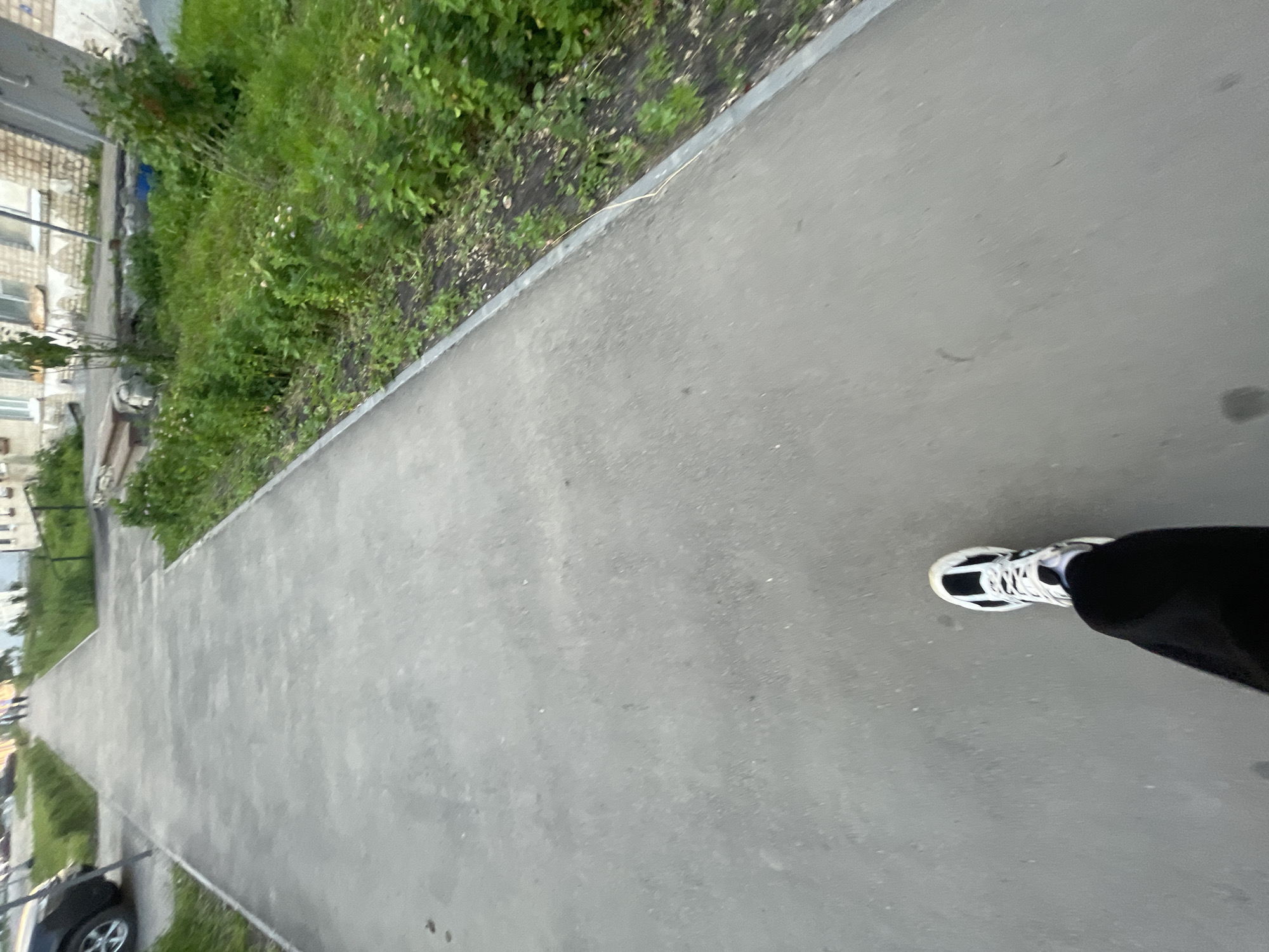 The image depicts a first-person perspective of someone walking on a paved sidewalk. The sidewalk is gray and appears to be in good condition, with a grassy area to the right. In the distance, there are buildings and some trees. A black car is visible on the left side of the image. The person is wearing black pants and white and black athletic shoes. The overall environment suggests a residential area in a city. Based on the context provided, this scene is located in the city of Krasnye Baki, Russia.