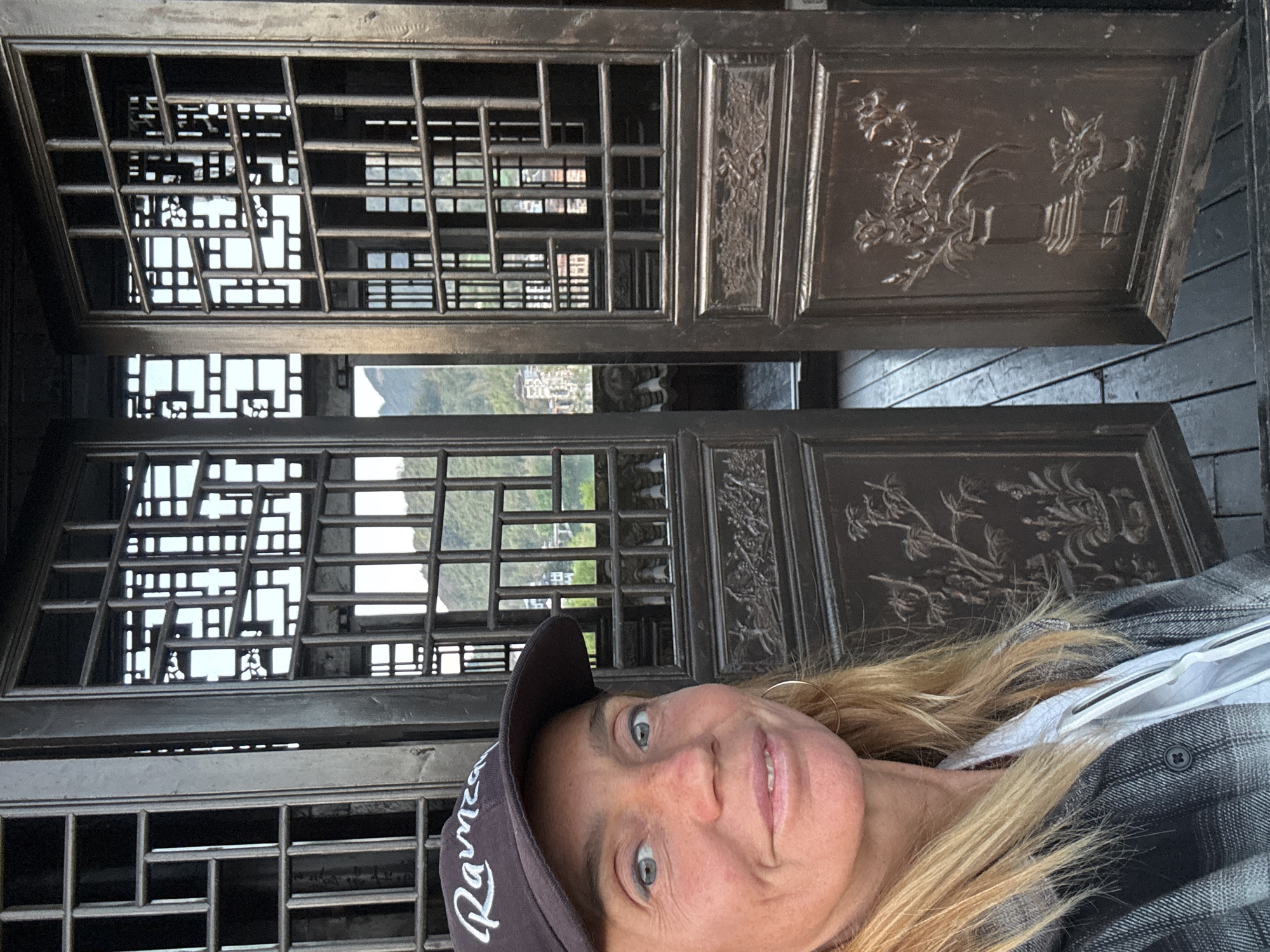 A medium shot depicts a woman in front of an ornate set of doors. The woman is wearing a baseball cap, a light-colored top, and a grey-and-black plaid shirt. Her blond hair is visible. The doors are dark and feature intricate latticework and carved panels. Through the openings in the latticework, a landscape with green hills is visible. The setting is Zhuoshui, China.