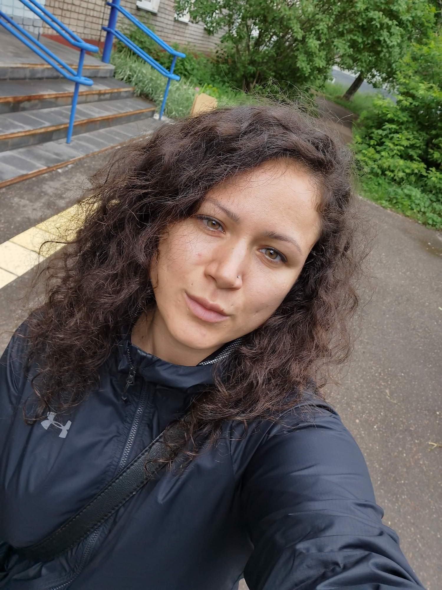 The image captures a head-and-shoulders, selfie-style portrait of a female subject outdoors. The woman has long, dark brown, curly hair that appears to have some volume and slight frizz. Her eyes are hazel or light brown, and a small silver stud piercing is visible on her left nostril. Her lips are naturally colored and appear full. She exhibits a neutral facial expression while looking directly at the camera. The subject is wearing a dark blue or black lightweight jacket, possibly a windbreaker, which is zipped up the front. A grey Under Armour logo is distinctly visible on the left chest area of the jacket. A dark, narrow strap, consistent with a shoulder bag, crosses her body diagonally from her left shoulder down towards her right hip.
The background on the left side of the image features a light-colored brick building with a series of weathered, possibly concrete or stone, steps leading up to an entrance. Blue metal railings are installed on both sides of these steps, guiding access. Green vegetation, including grass and bushes, is present around the base of the steps and adjacent to the building. On the ground directly in front of the subject, extending to the left, is a yellow tactile paving strip.
The background on the right side of the image shows a paved, asphalt surface, likely a walkway or road, which appears somewhat damp. Scattered green foliage, including bushes and trees, lines this pathway. A distant tree with a slight lean is visible further into the background. The overall illumination suggests natural daylight, possibly under overcast conditions, as indicated by the soft lighting and lack of harsh shadows. The environment is consistent with an urban or park-like setting.