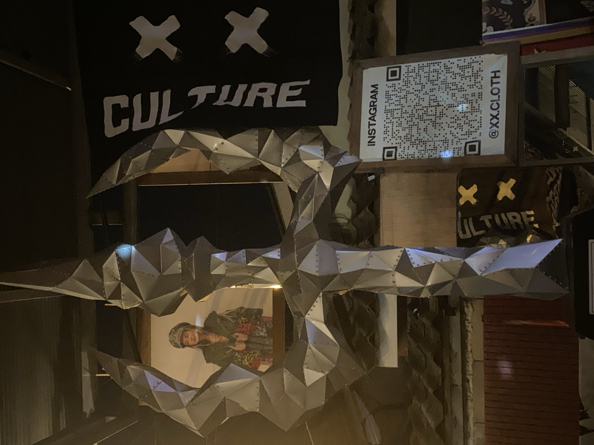 The image depicts an installation in Thong Sala, Thailand, featuring a metallic sculpture resembling a stylized skull or mask. A banner with the word "CULTURE" in white block letters is visible in the background, adorned with two stylized "X" marks. A QR code labeled "INSTAGRAM @XX.CLOTH" is affixed to a wooden structure adjacent to the banner. The sculpture frames a partial view of a person wearing patterned clothing and headwear. The environment appears to be a dimly lit, possibly urban or commercial space.