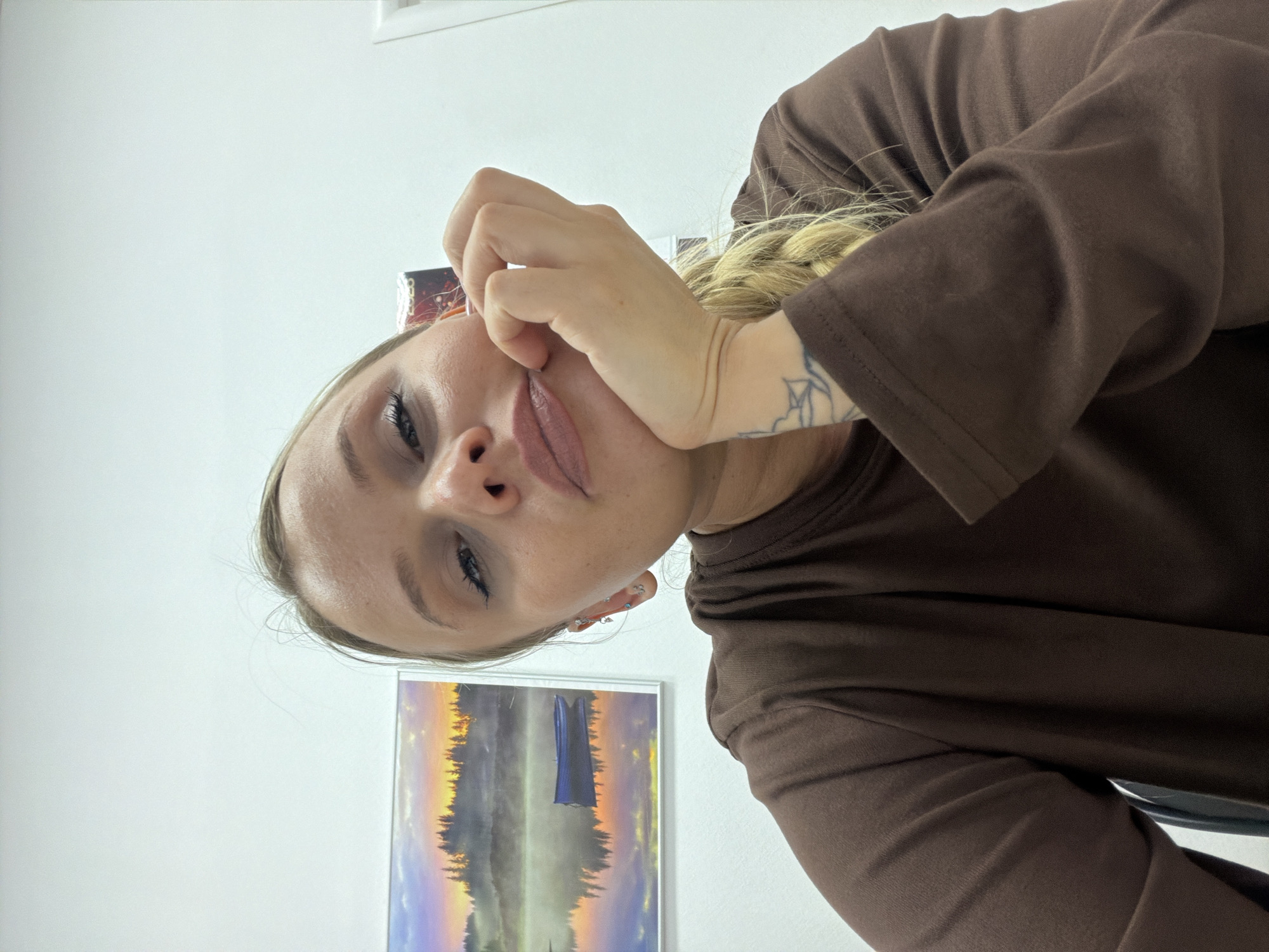 A fair-skinned young woman with blonde hair in a braid is positioned in the center of the frame, tilting her head to the left. She is wearing a brown long-sleeved shirt and has a tattoo visible on her right inner arm. Her right hand is near her face, with her fingers touching her cheek. The background is a white wall. A framed picture depicting a landscape with trees and a sunset is on the wall behind her. The location of Sarapul, Russia, cannot be confirmed based on the provided image.