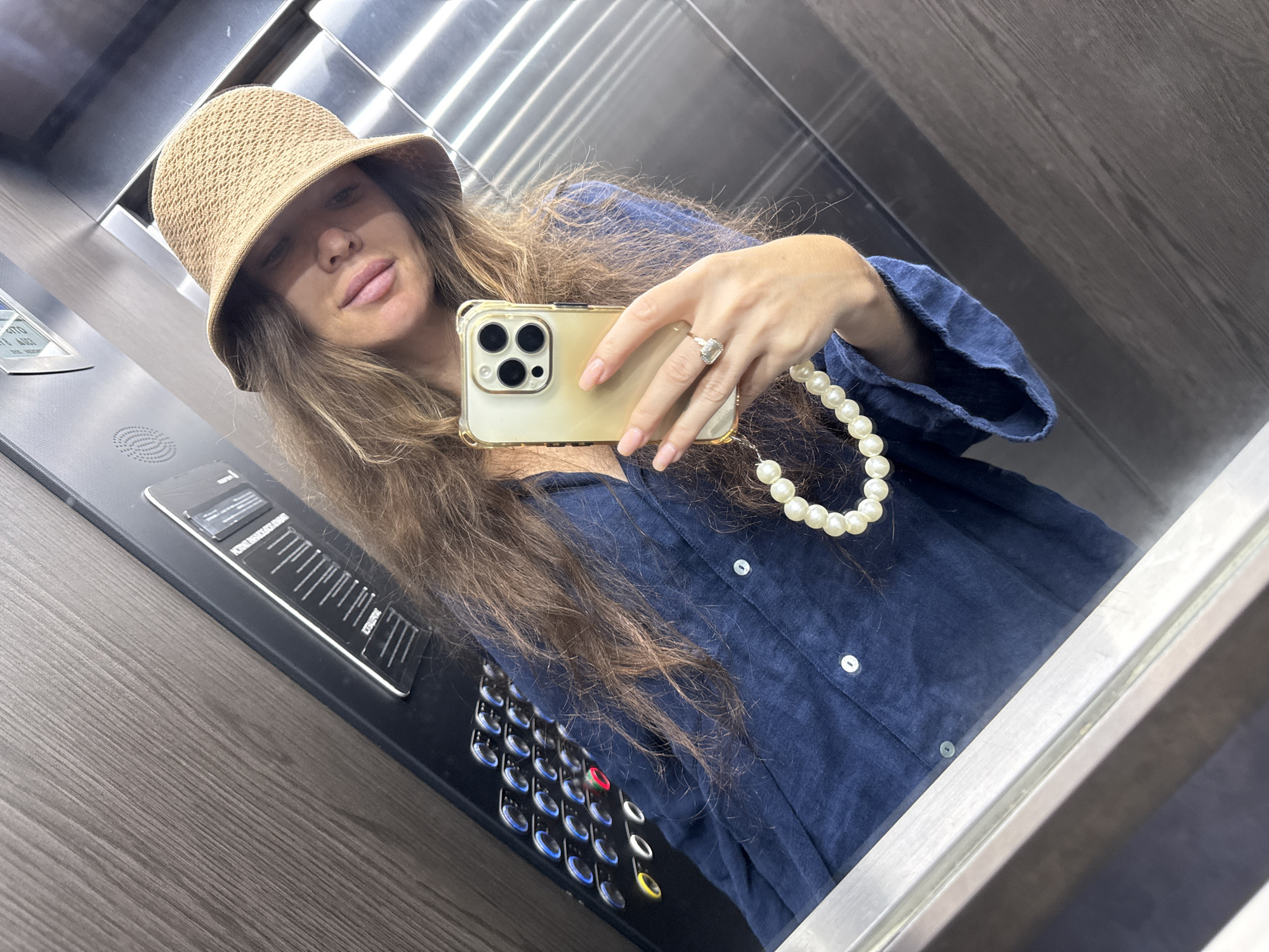 A woman is taking a selfie in an elevator. She is wearing a straw hat, a navy blue shirt, and a pearl necklace. She is holding a gold iPhone in her right hand. The elevator has wood-paneled walls and a control panel with illuminated buttons. The image appears to have been taken in the city of Khimki, Russia.