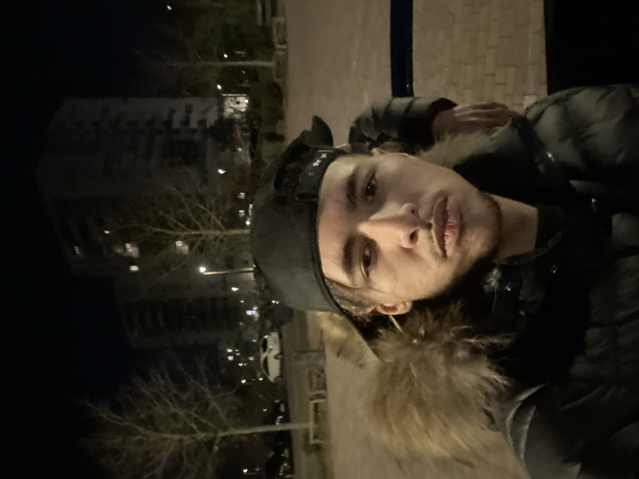 The image displays a nighttime urban scene in Ostwald, France. In the foreground, a male individual, appearing to be in his late teens or early twenties, with light brown skin, a thin mustache, and dark hair, faces directly towards the camera. He wears a black baseball cap backward and a black puffer jacket with a prominent light brown fur-lined hood. A partial view of a second individual, including an arm and shoulder dressed in a dark jacket, is visible behind the primary subject on the right side of the frame. The background consists of a paved ground area, several bare deciduous trees, and multiple illuminated streetlights. Two dark, multi-story residential buildings with illuminated windows are visible in the distance. Multiple vehicles, including a white car, are parked further in the background. A blue horizontal barrier is partially visible on the right.
