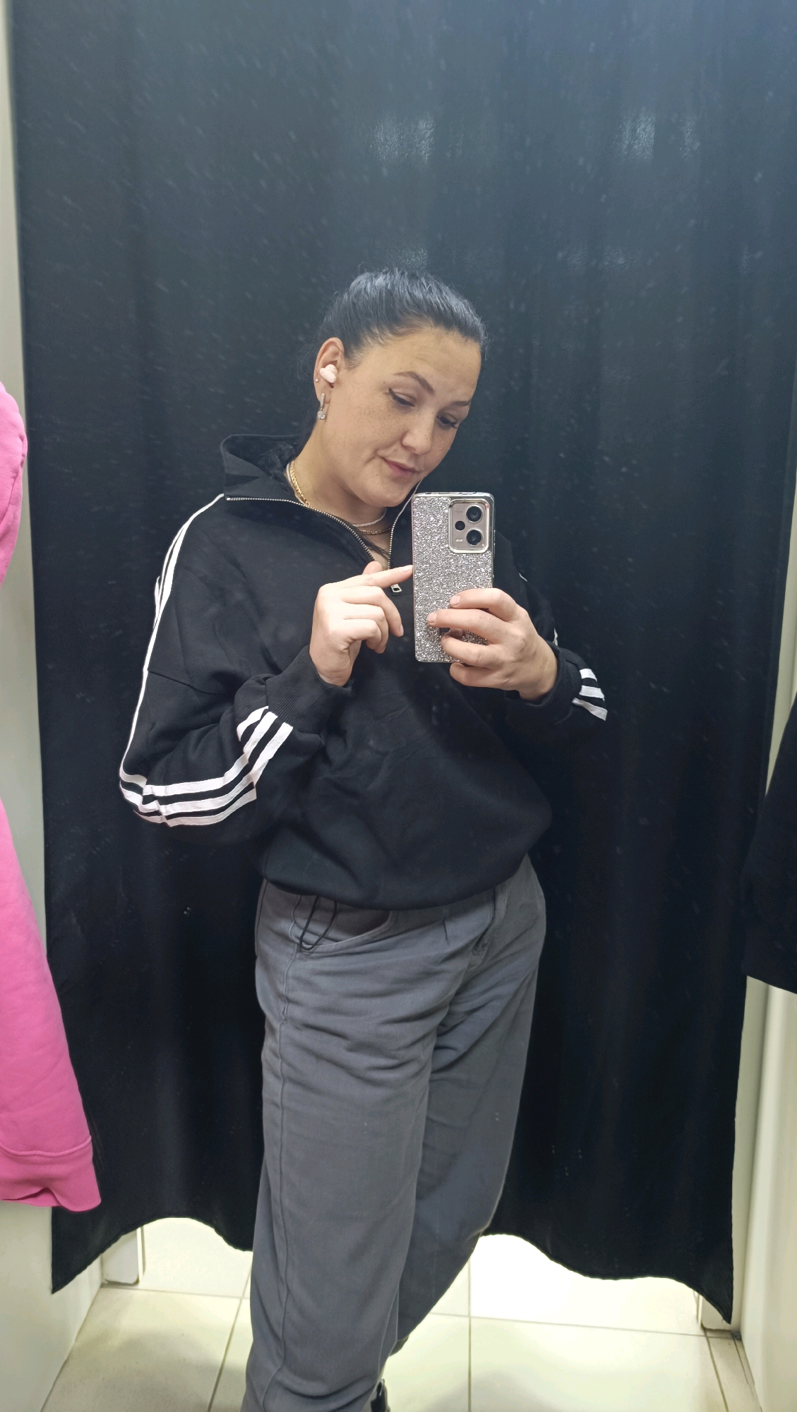 A woman is standing in a fitting room, taking a mirror selfie. She is wearing a black hooded sweatshirt with white stripes on the sleeves and grey pants. She has her hair pulled back in a bun and is wearing white earbuds. She is holding a silver, glitter-covered phone in her right hand. To her left, a pink garment is hanging on a rack. The fitting room has black curtains and a tiled floor.