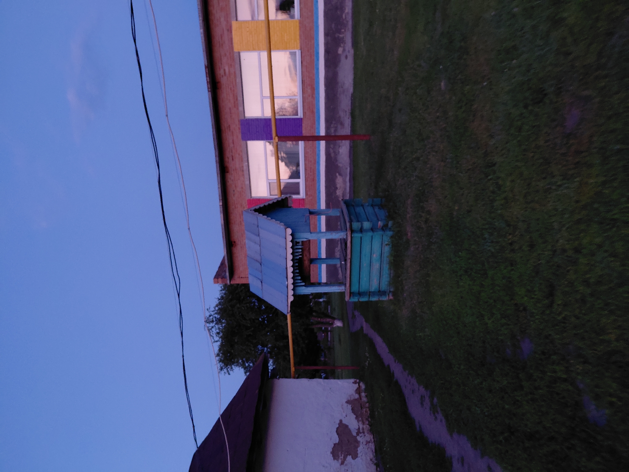 The image depicts an outdoor scene with a twilight sky. In the foreground, a small, weathered, blue and green wooden well stands on a patch of grass. To the left, a white building with peeling paint is visible, with a dark-pitched roof. A narrow, winding path, possibly made of stone or concrete, leads from the building towards the well. Behind the well and building, a red brick structure with multiple windows is situated. These windows have colorful rectangular accents in purple and yellow. The building also has a horizontal band of blue and white paint near its base. A yellow pipe runs horizontally across the mid-ground, supported by red posts, extending from the white building towards the brick structure. Electrical wires are strung across the upper portion of the image, against the blue sky. The overall environment appears to be a rural or suburban setting, possibly a schoolyard or a communal area.