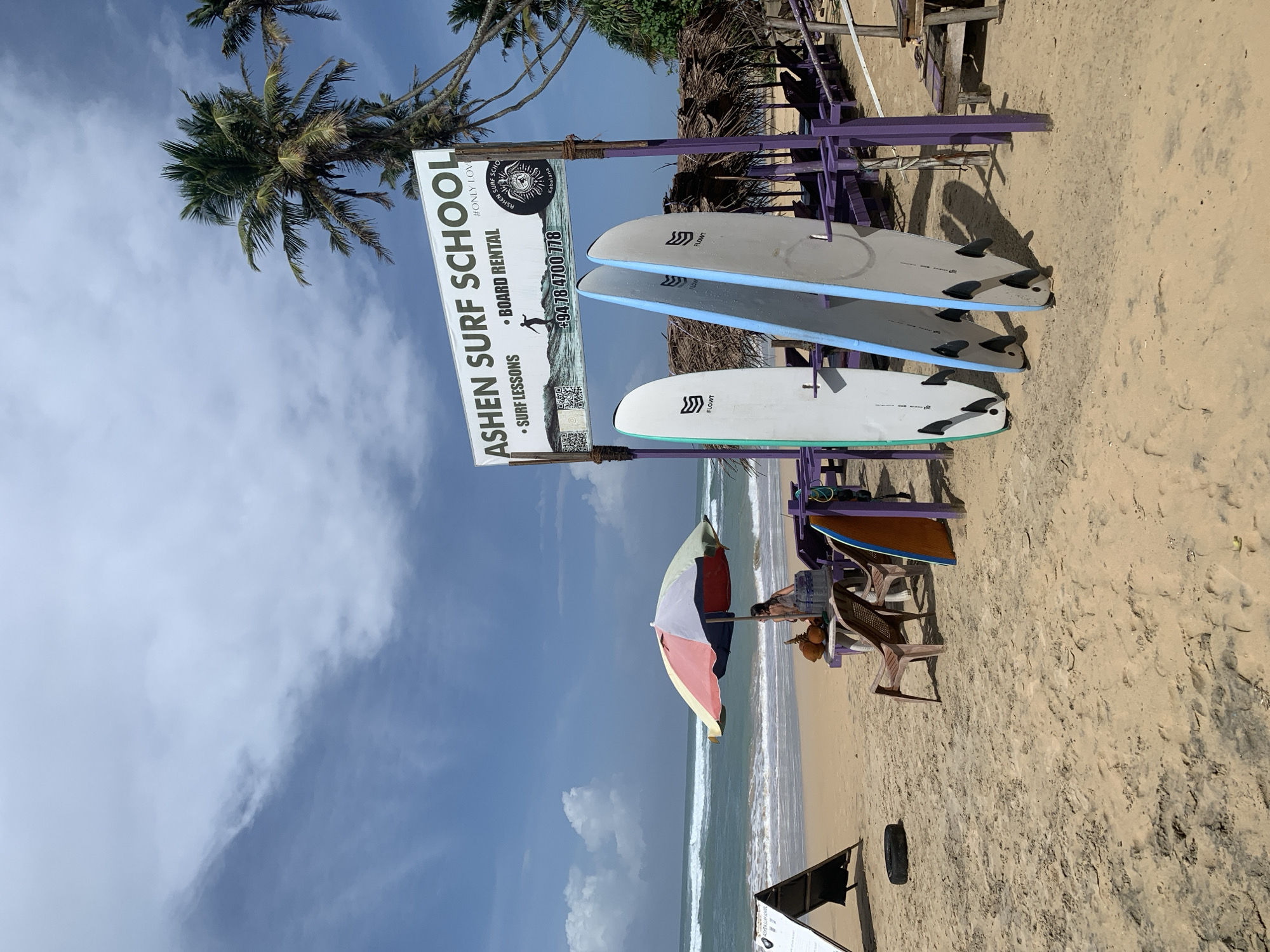 This is a ground-level, eye-level, medium shot of a beach scene in Ahangama, Sri Lanka, under a partly cloudy sky. In the foreground, three surfboards are propped up in the sand. Two are light gray with blue trim, and one is orange. Behind them, a purple wooden structure with thatched roofs provides shade for several lounge chairs and tables. A woman sits on a chair under a colorful umbrella near the water's edge. In the background, waves crash onto the sandy shore, and palm trees line the coast. A sign reads "ASHEN SURF SCHOOL," listing surf lessons and board rental, along with a phone number and social media QR codes.